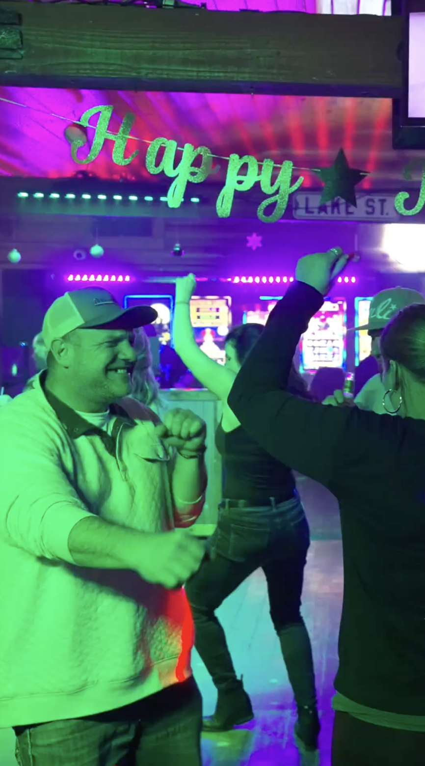 People dancing and enjoying a party under colorful lights, with a festive sign that says 'Happy' and holiday decorations in the background.