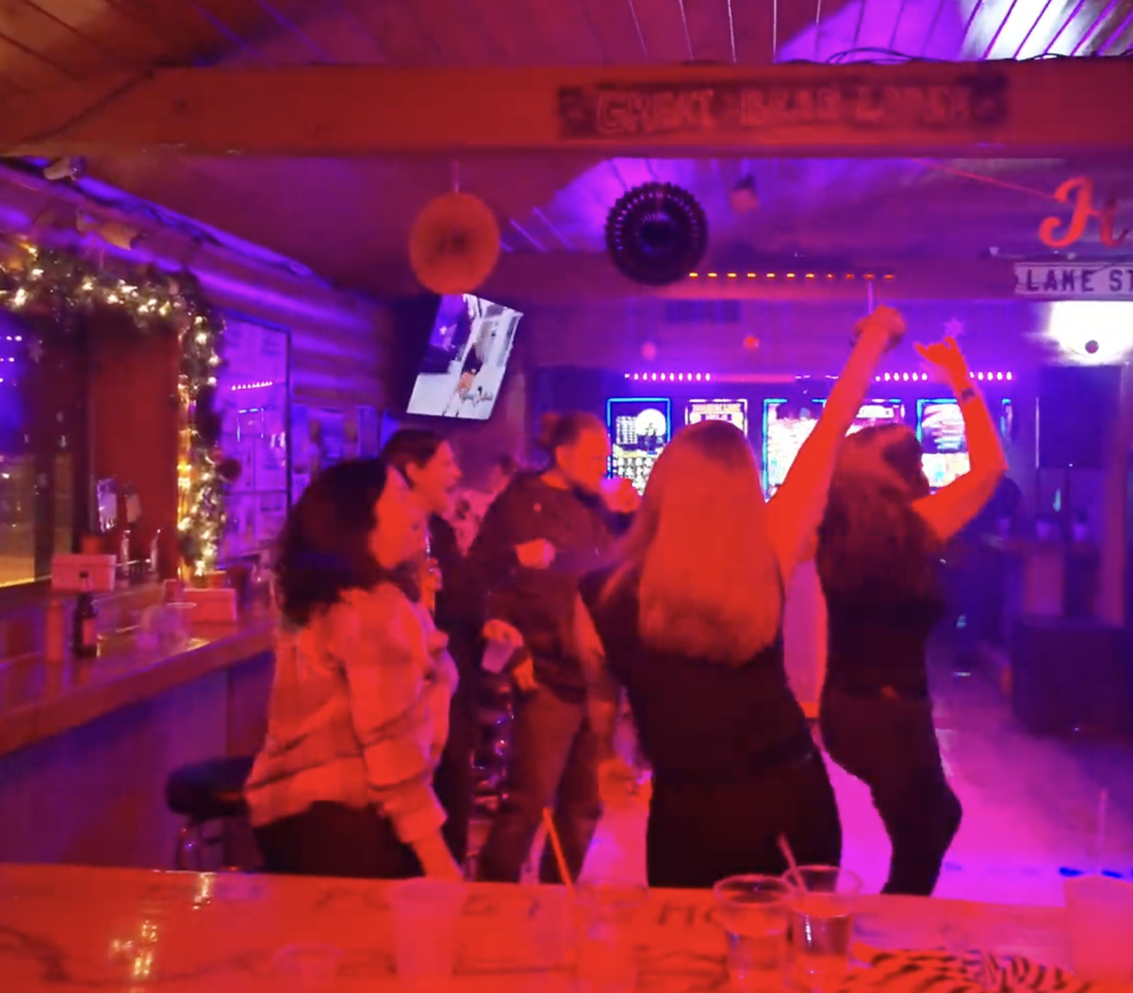 People at a bar dancing and socializing under colorful purple and red lighting with neon signs and a television screen in the background.