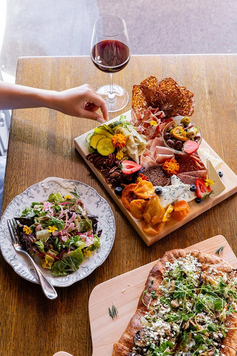 A table with a charcuterie board, a glass of red wine, a salad in a decorative bowl, and a pizza. A person is reaching for a piece of the charcuterie board.