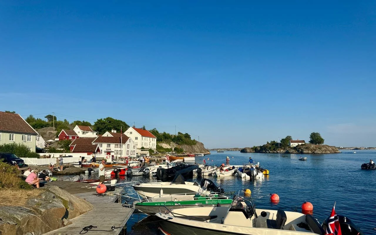 A scenic harbor with boats docked along a pier, houses on a hillside, and clear blue sky.