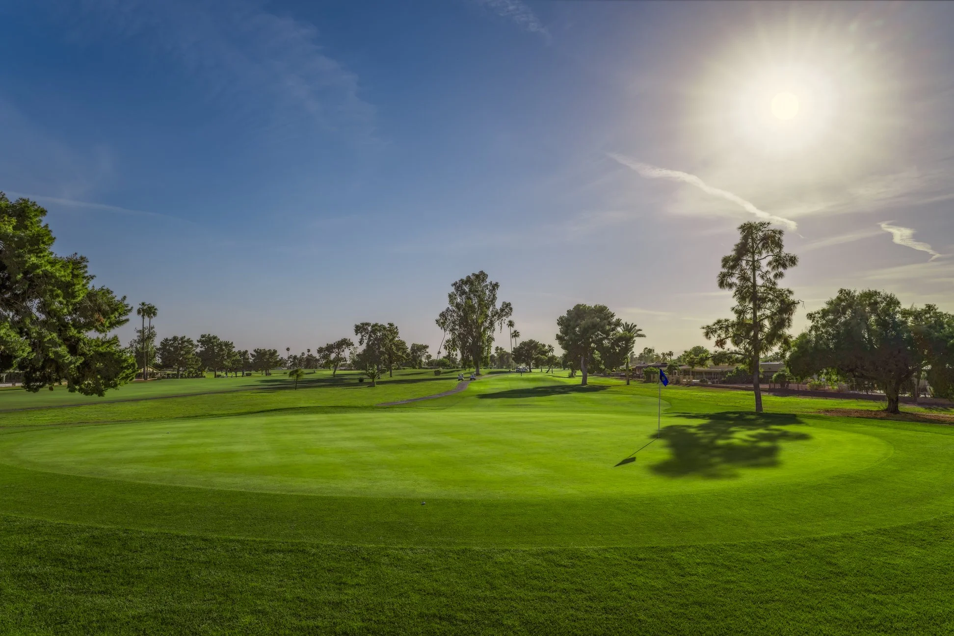 A golf course with green grass, trees, a flag on the putting green, and a bright sun in a mostly clear sky. View of the 7th hole at Palmbrook Golf Club in Sun City, Arizona.