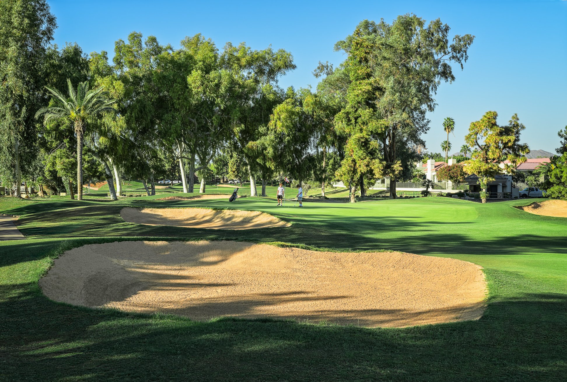 A golf course with a sand trap in the foreground, lush green grass, tall trees, and two people playing golf in the distance under a clear blue sky on the 1st hole of Scottsdale Country Club golf course in Scottsdale Arizona.