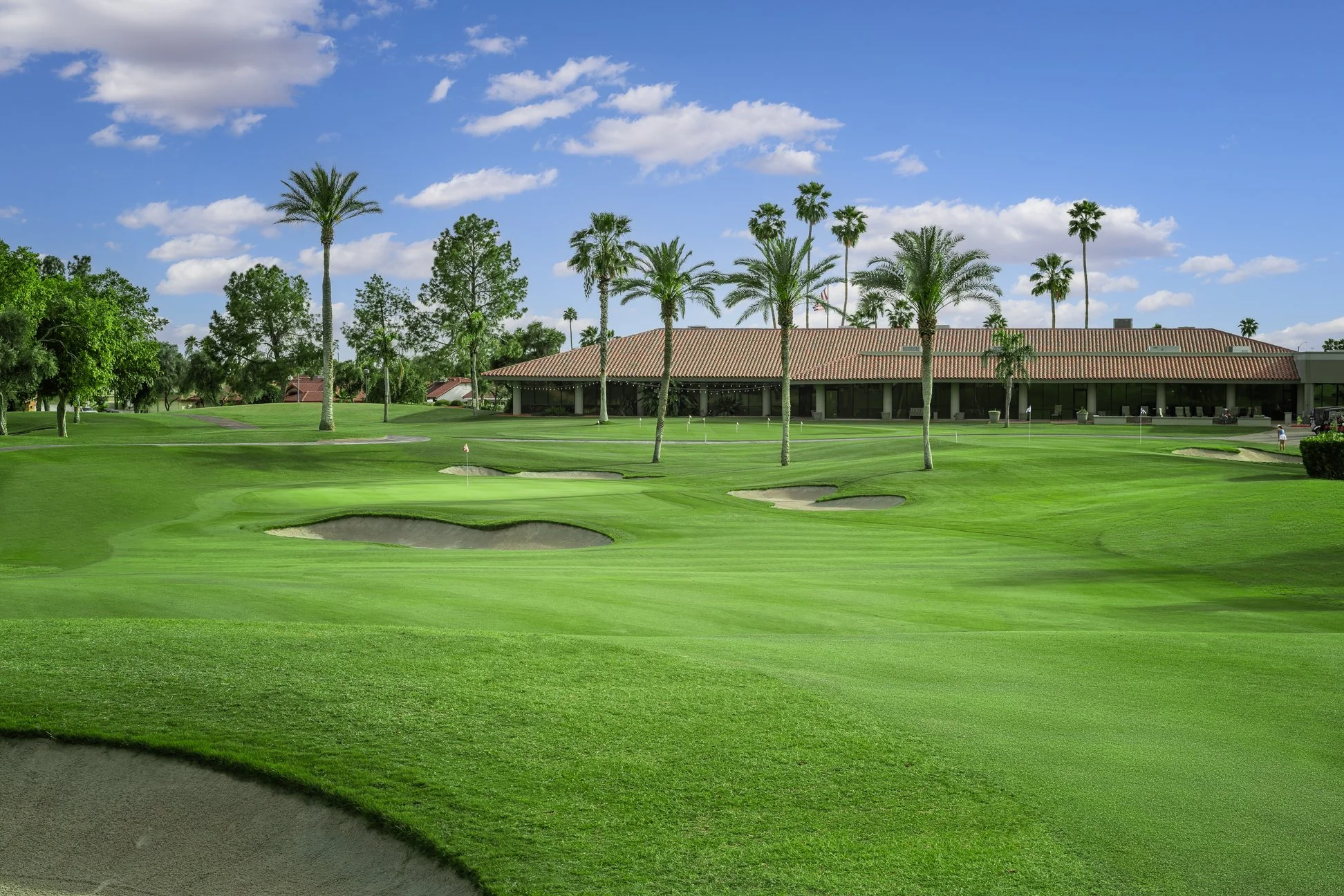 A green golf course with sand bunkers, a building with a tiled roof, and tall palm trees under a partly cloudy blue sky. View of the clubhouse from the fairway at Briarwood Country Club in Sun City West Arizona.