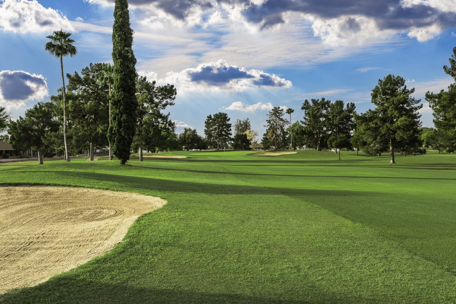 A golf course with lush green grass, a sand trap in the foreground, numerous trees including palms and cypress, and a partly cloudy sky, showing the 13th hole at Union Hills Golf Club in Sun City, Arizona.