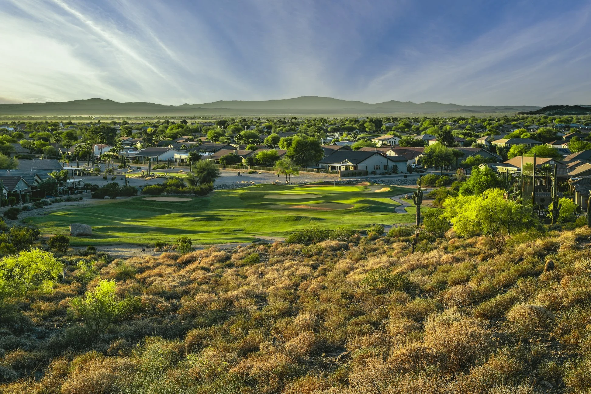 A landscape of a suburban neighborhood with houses, trees, and a golf course, with mountains in the background and a blue sky with wispy clouds at San Tan Highlands Golf Club, Arizona.