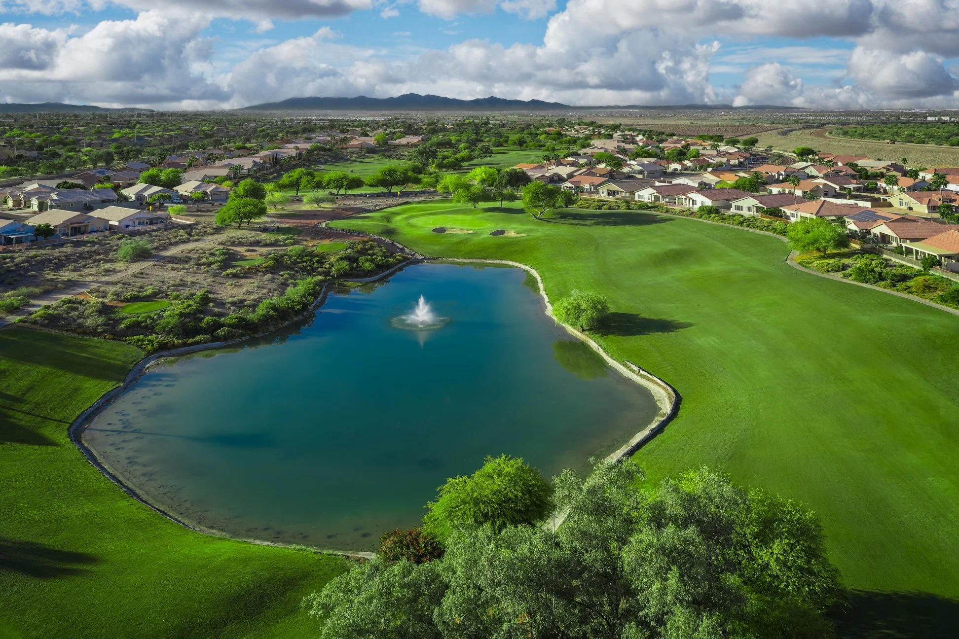 A scenic view of a golf course with green grass, a pond with a fountain, surrounding trees, residential houses, and mountains in the background under a partly cloudy sky.