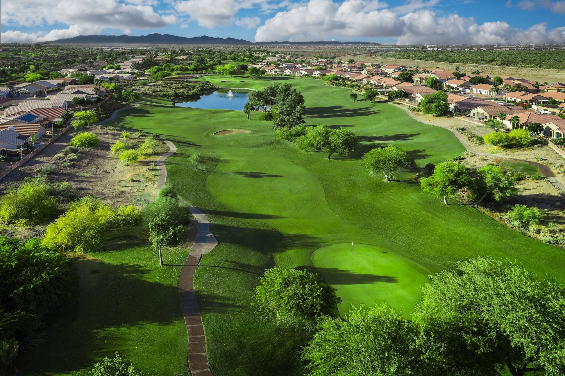 Aerial view of a golf course with green fairways, sand traps, trees, water features, and a flag, surrounded by residential houses under a partly cloudy sky at Coyote Lakes Golf Club in Surprise, Arizona.