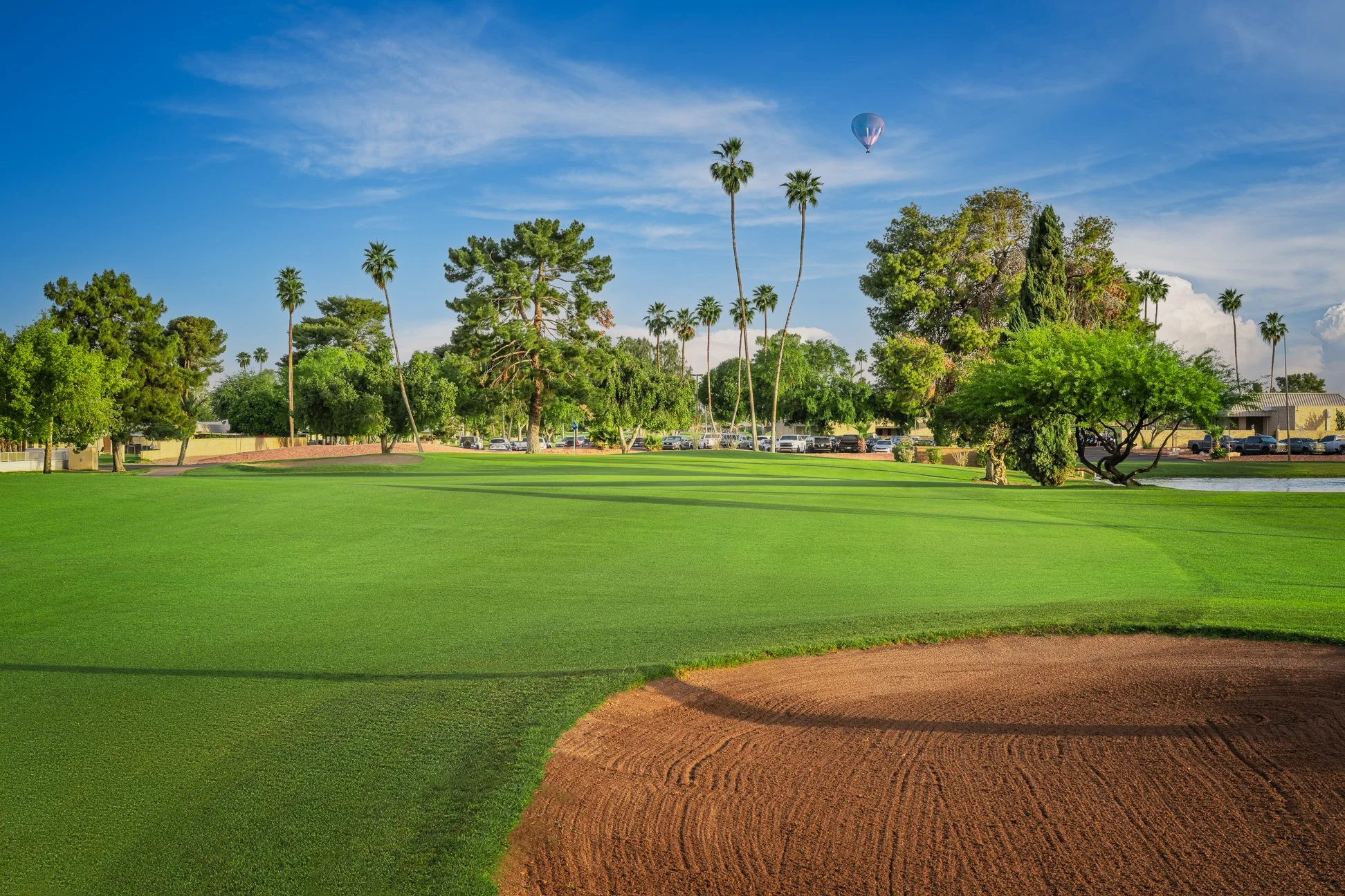 Golf course with green grass, sand trap in foreground, trees, parked cars, and a hot air balloon in a blue sky with some clouds. View from the 18th hole at Palmbrook Golf Club in Sun City, Arizona.