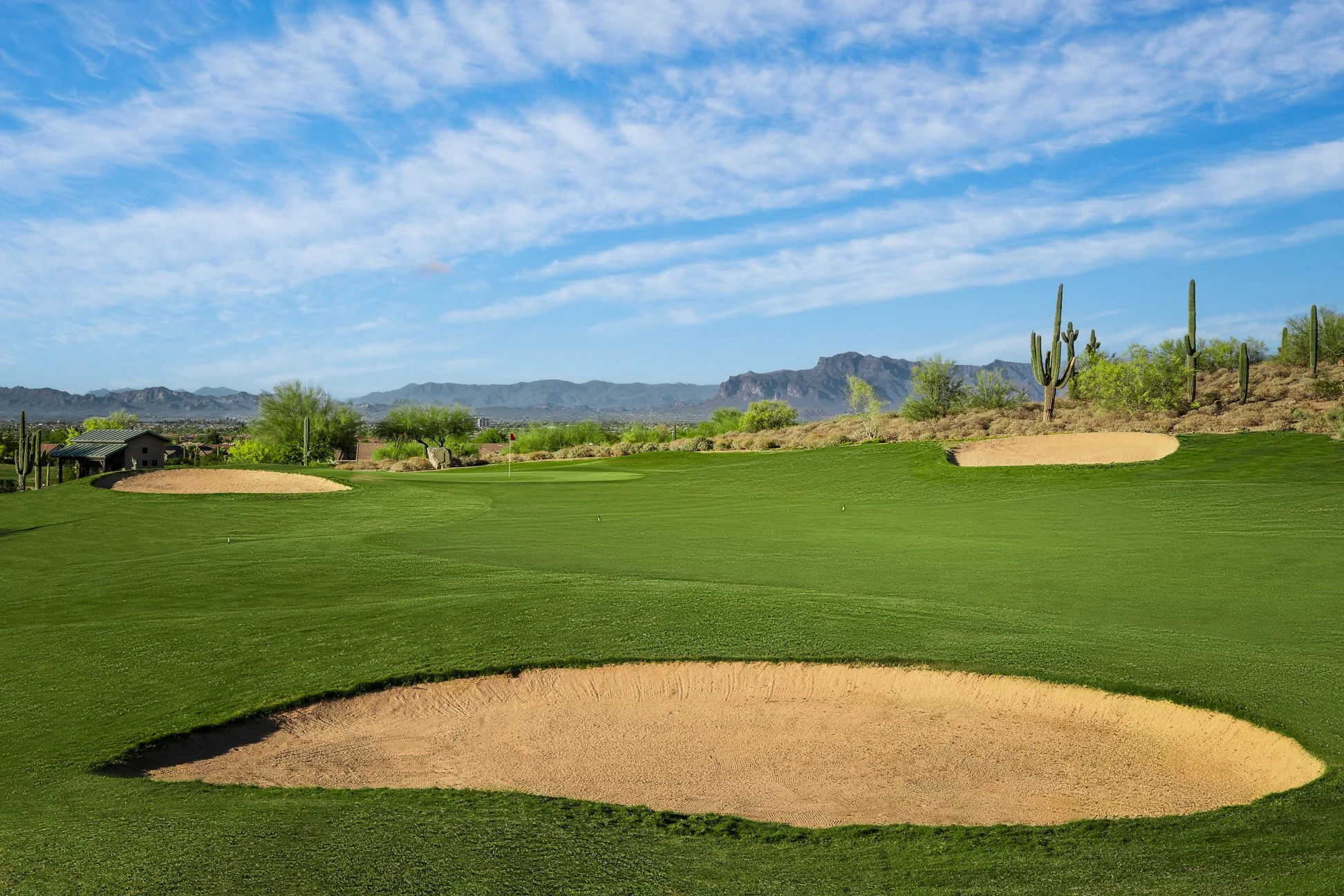 A golf course with green fairways, sand bunkers, a flag indicating the hole's location, along with cacti, desert shrubs, and mountains in the background under a blue sky with scattered clouds at San Tan Highlands Golf Club, Arizona