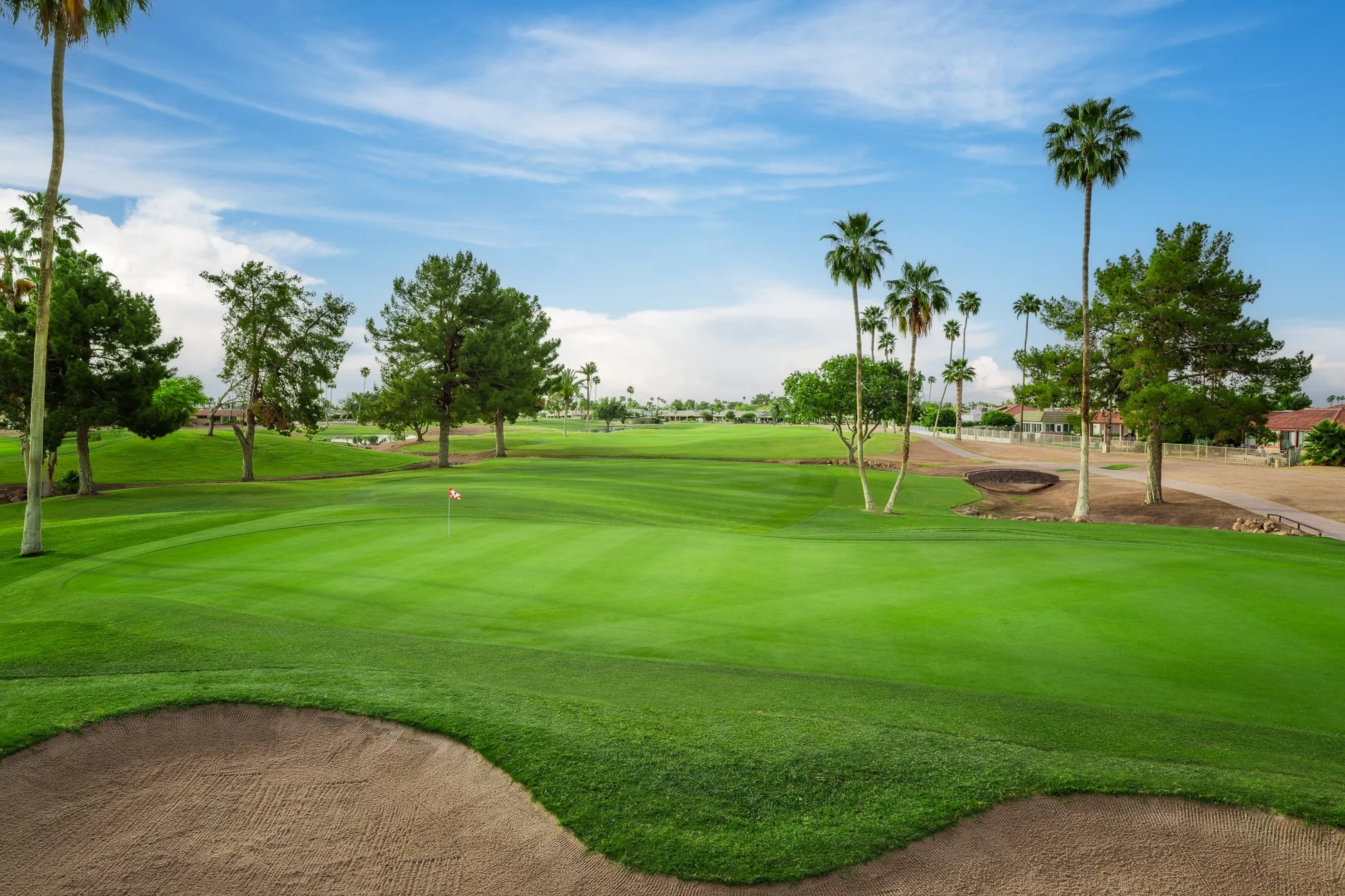 A golf course with lush green grass, sand bunkers, and tall palm trees under a partly cloudy blue sky on the 15th hole at Briarwood Country Club in Sun City West, Arizona.
