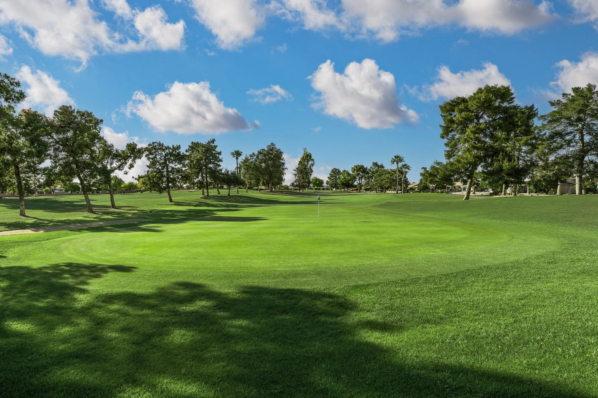 A golf course with lush green grass, a flag in the distance, surrounded by trees, under a blue sky with scattered white clouds. View of the 5th hole at Union Hills Golf Club in Sun City, Arizona,