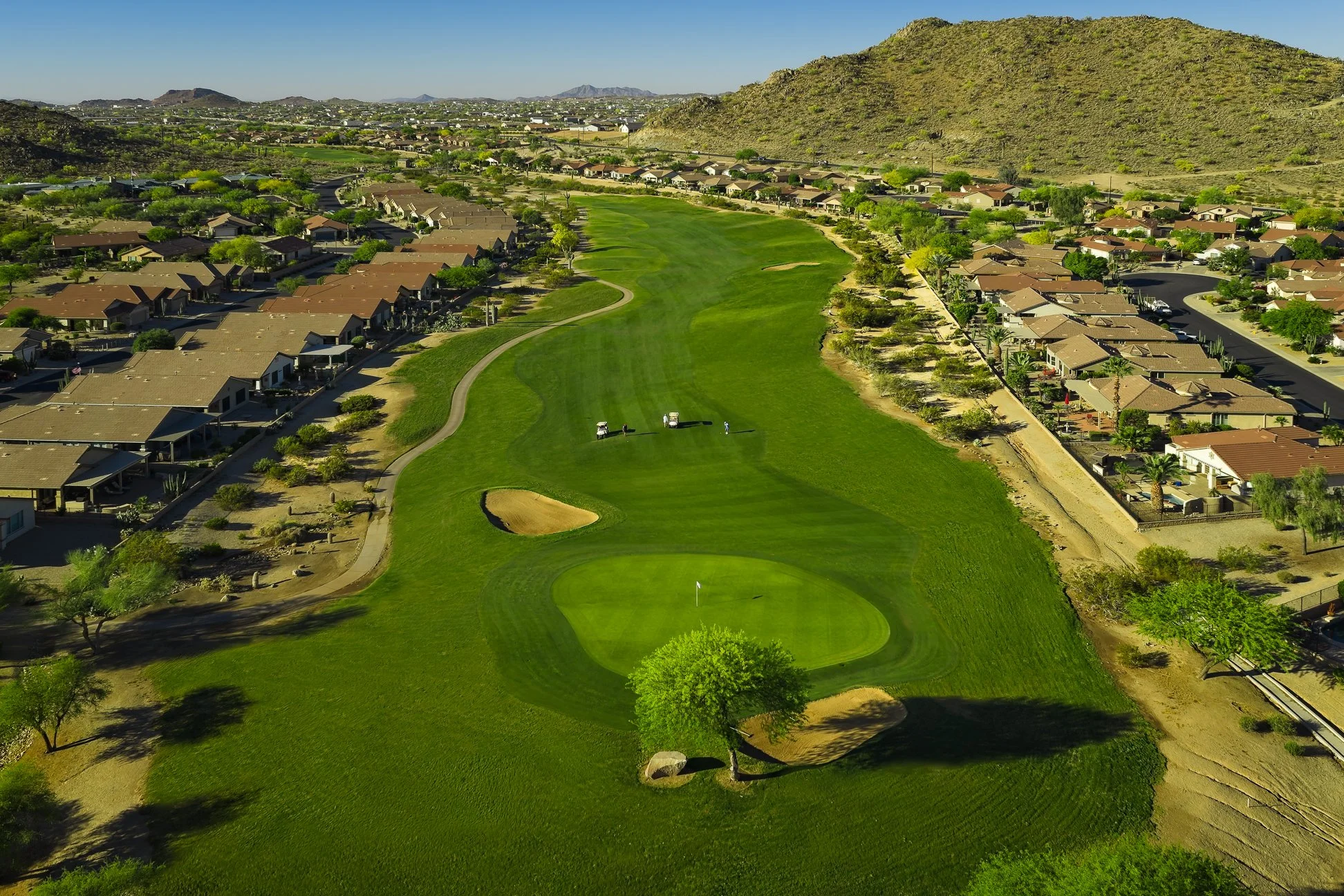 An aerial view of a golf course with a green, sand trap, and pathways, surrounded by suburban houses and desert landscape, with mountains in the background.