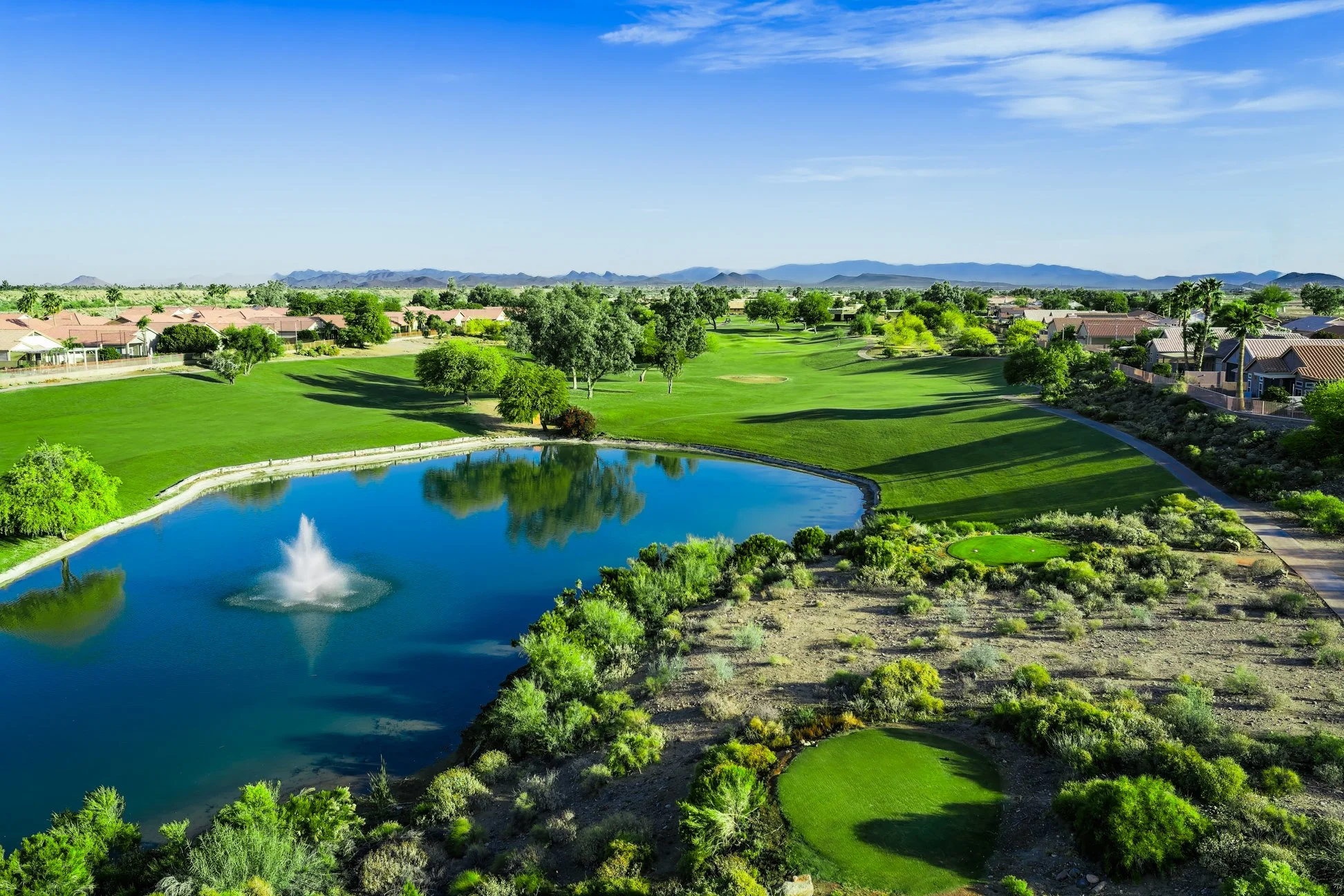 A view of a golf course with a pond and fountain, surrounded by green grass, trees, and residential houses under a blue sky with some clouds, with mountains in the background at Coyote Lakes Golf Club in Surprise, Arizona.