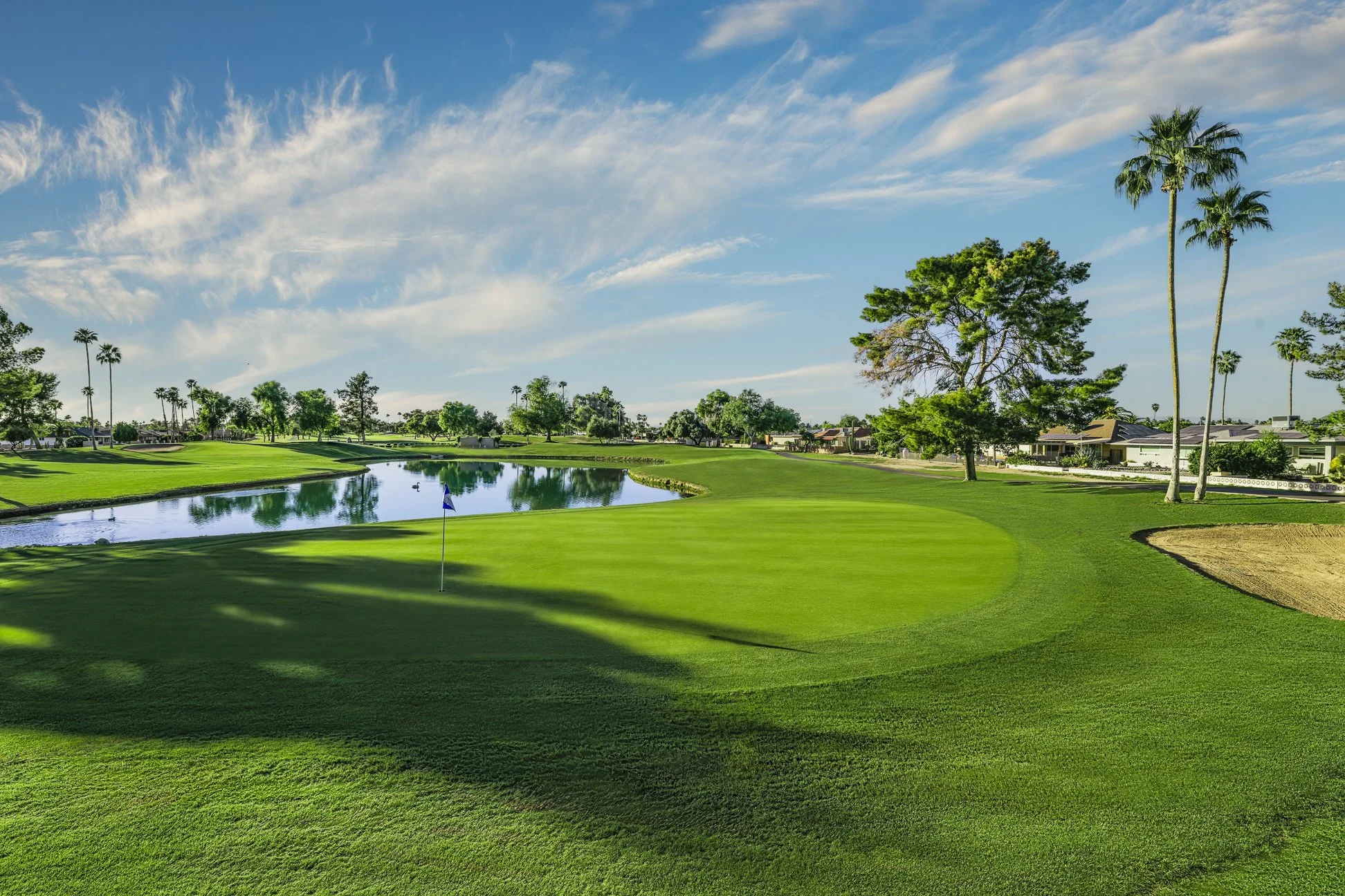 A golf course with a green and a flag near a water hazard, surrounded by palm trees and houses under a partly cloudy sky. View of the 12th hole at Union Hills Golf Club in Sun City, Arizona.