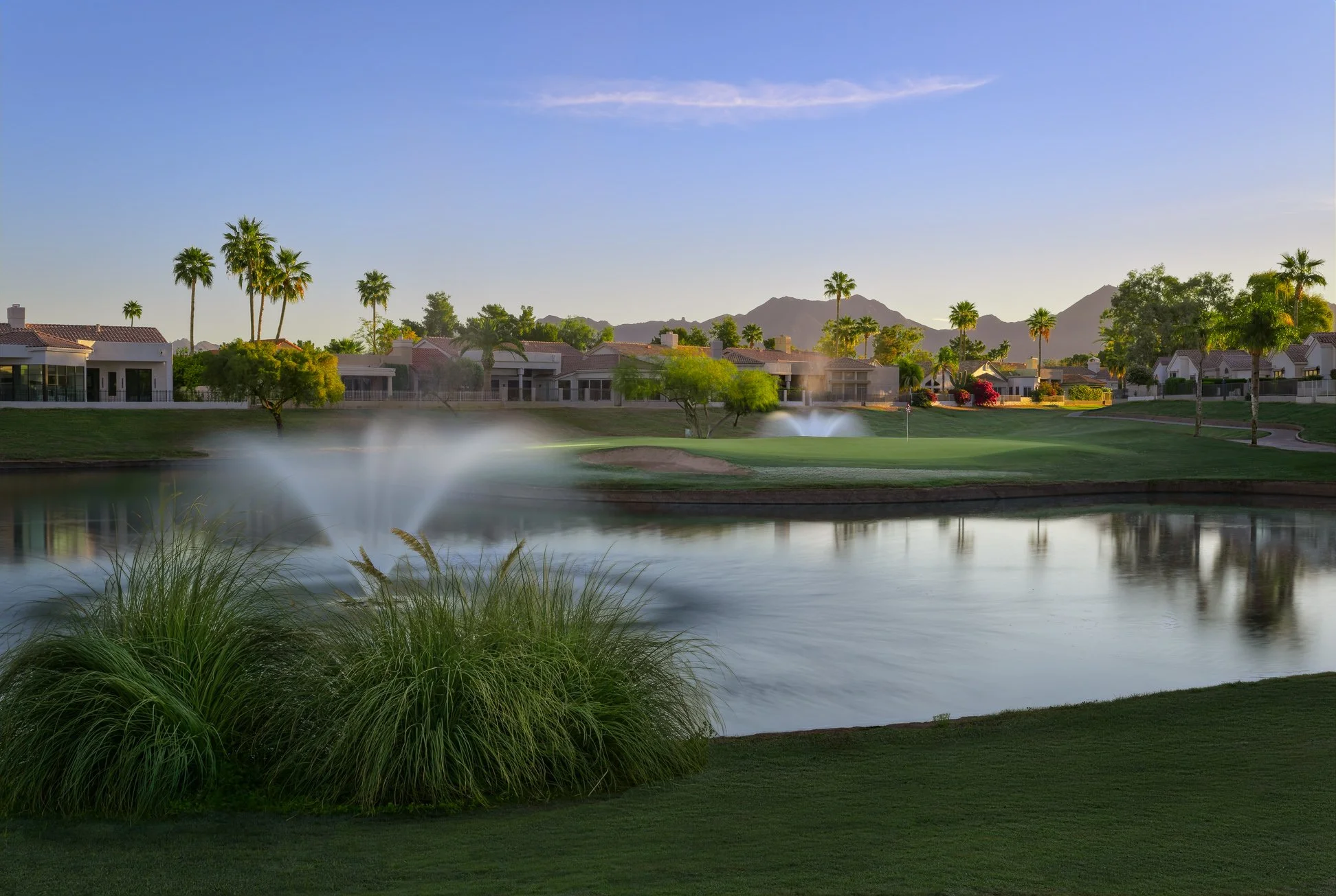 A scenic view of a golf course with green grass, a pond with a fountain, and residential houses with palm trees in the background, mountains in the distance, under a clear blue sky at Scottsdale Country club in Scottsdale Arizona.