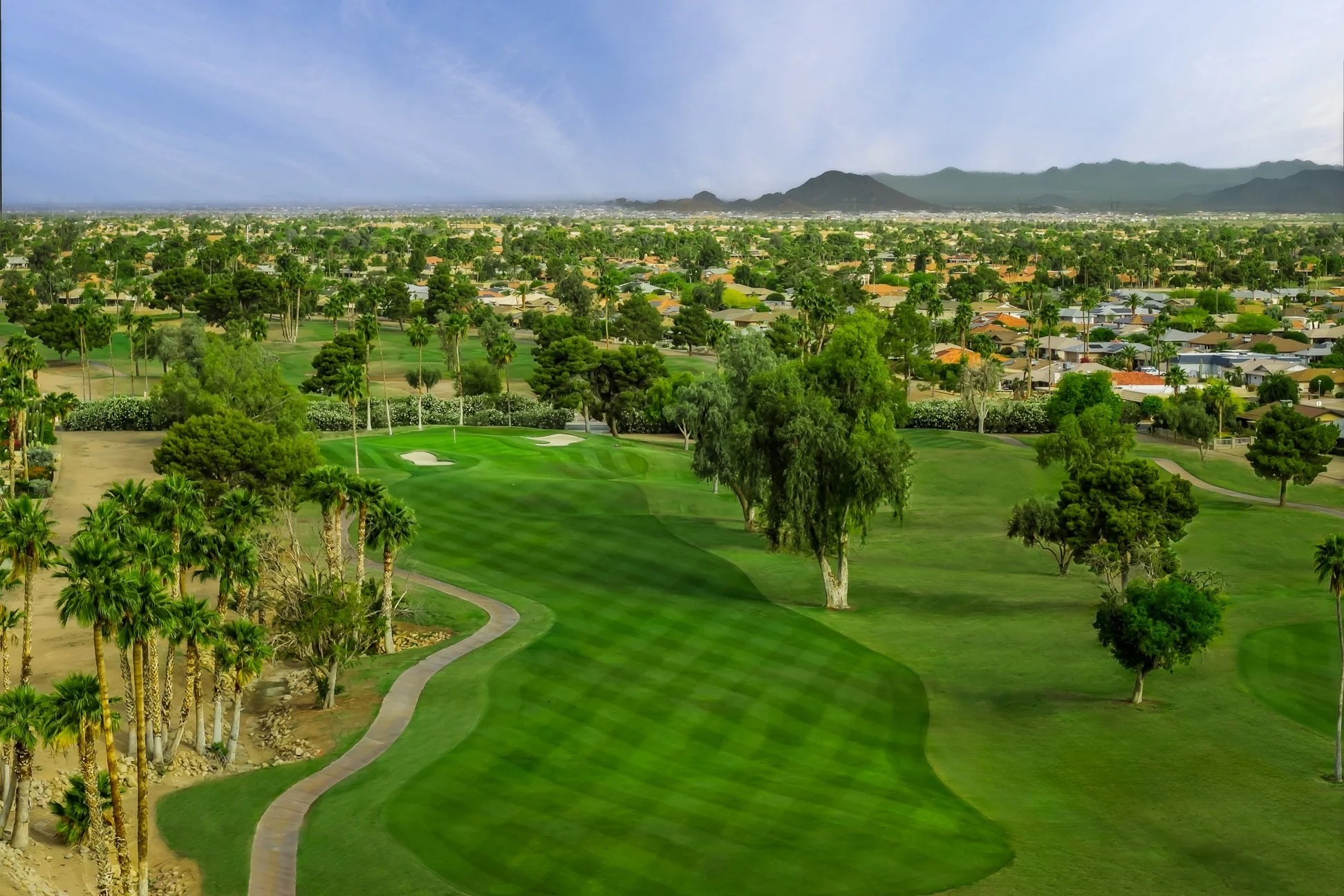 A golf course with lush green fairways and trees, neighboring residential houses, and mountains in the distance under a partly cloudy sky. Aerial view of Briarwood Country Club in Sun City West, Arizona.