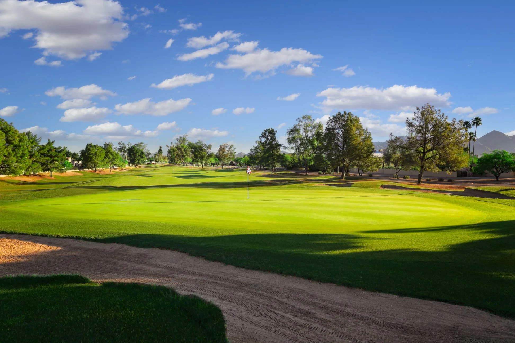 A golf course with green grass and a flag on the hole, surrounded by trees, mountains in the background, and a blue sky with scattered clouds. View of the 7th hole at Scottsdale Country Club in Scottsdale Arizona.