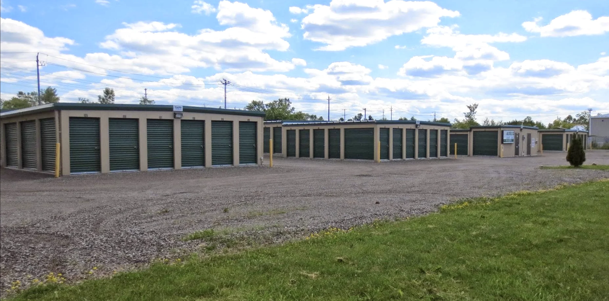 A storage facility with multiple storage units, each secured with a roll-up door, set against a partly cloudy sky and power lines, with a gravel lot and green grass in the foreground.