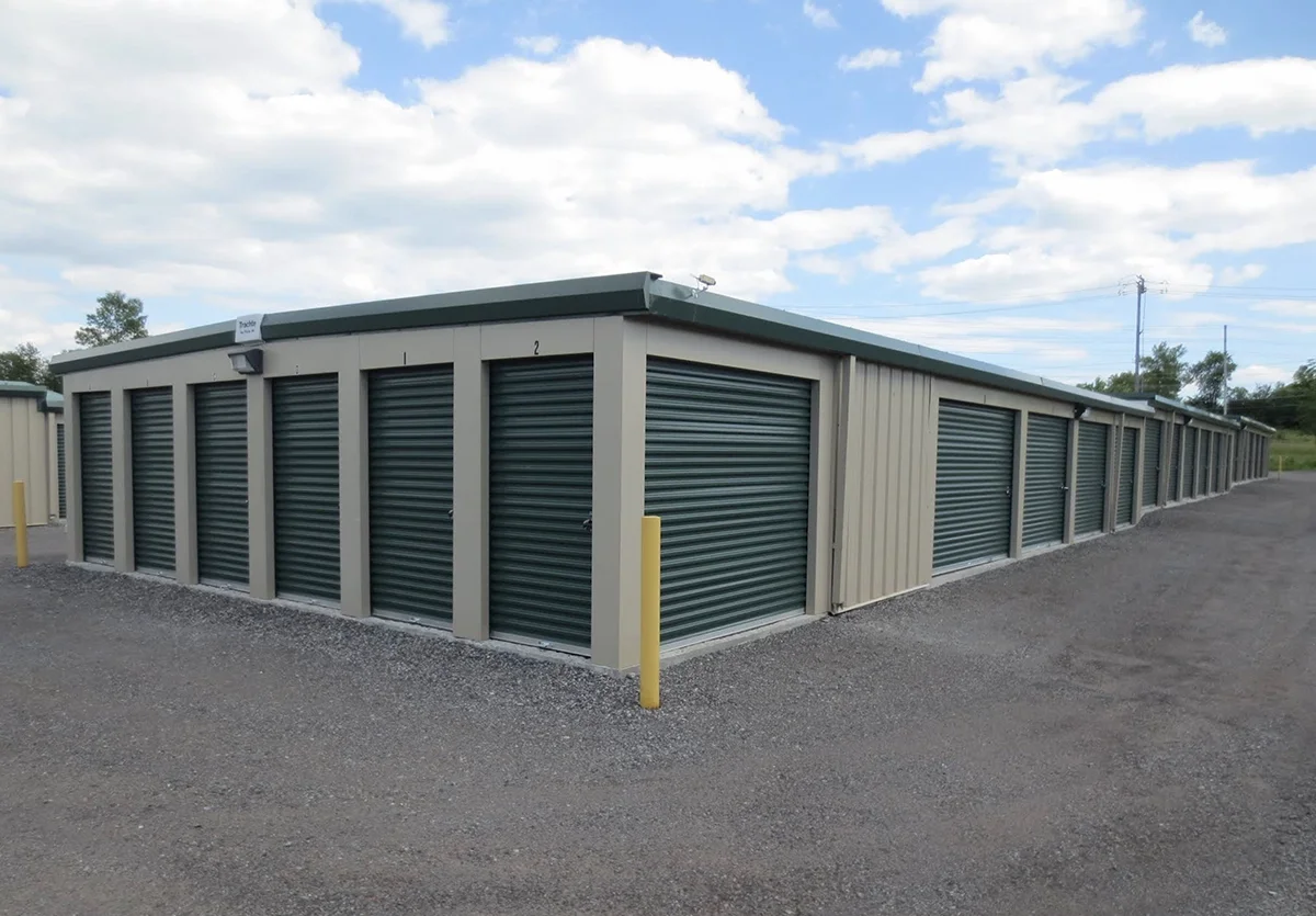 Exterior view of a row of storage units with green roll-up doors, set on a gravel lot under a partly cloudy sky.