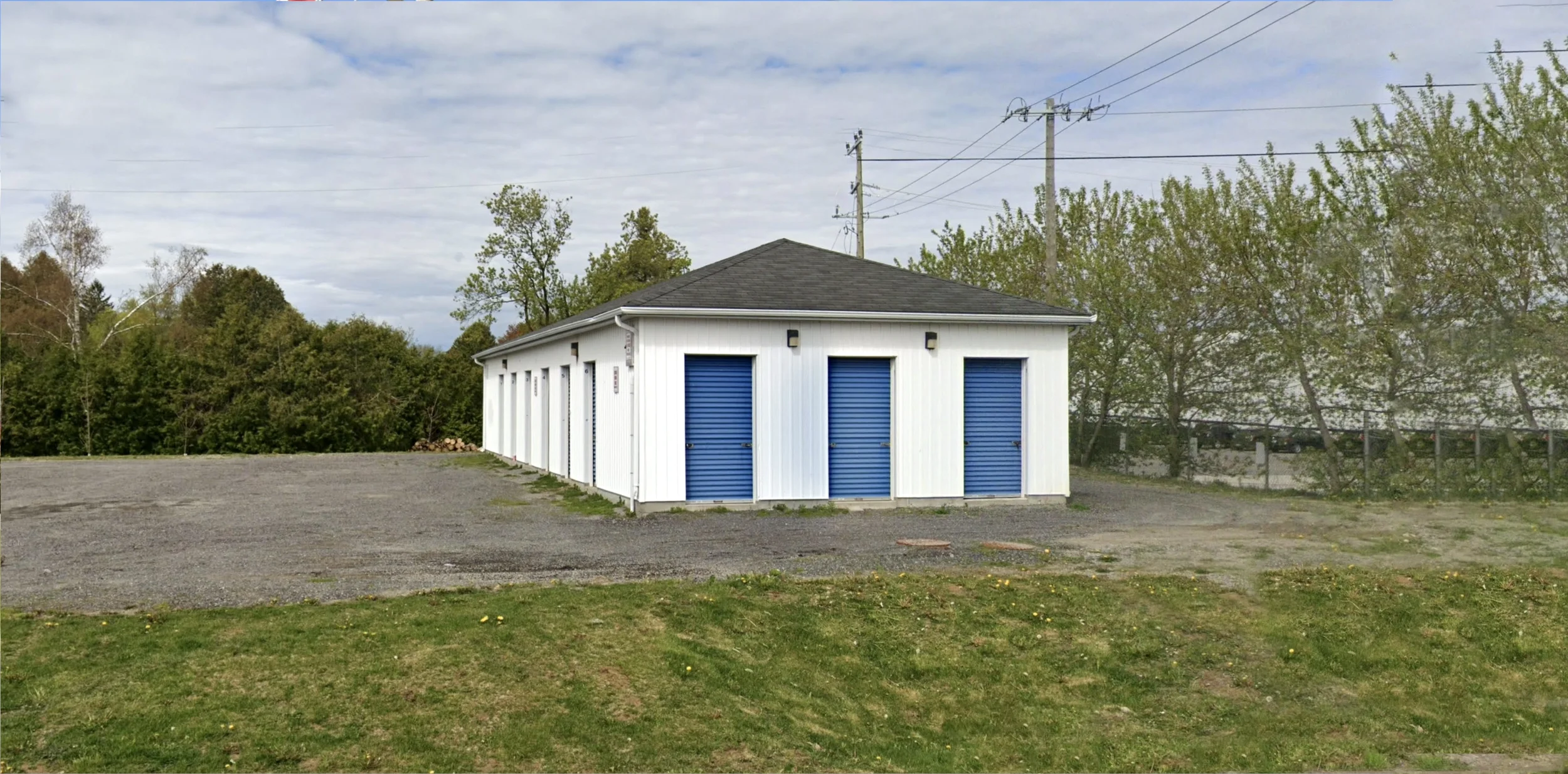 Small white building with blue garage doors and a dark gray roof, situated on a gravel lot with some grass in the foreground. Trees and power lines are in the background, under a partly cloudy sky.