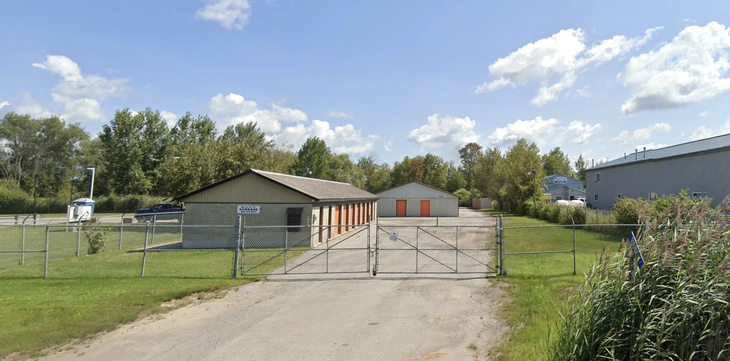 Fenced industrial storage area with small storage buildings, a gate, and a parking lot, surrounded by trees and a blue sky with clouds.
