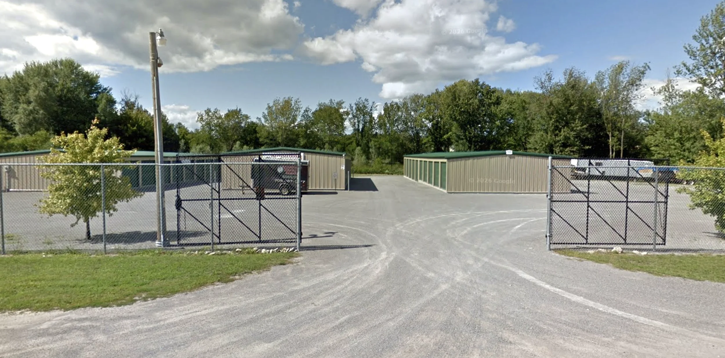 Self serve storage units enclosed by a chain-link fence, with two storage sheds, trees, and a partly cloudy sky.
