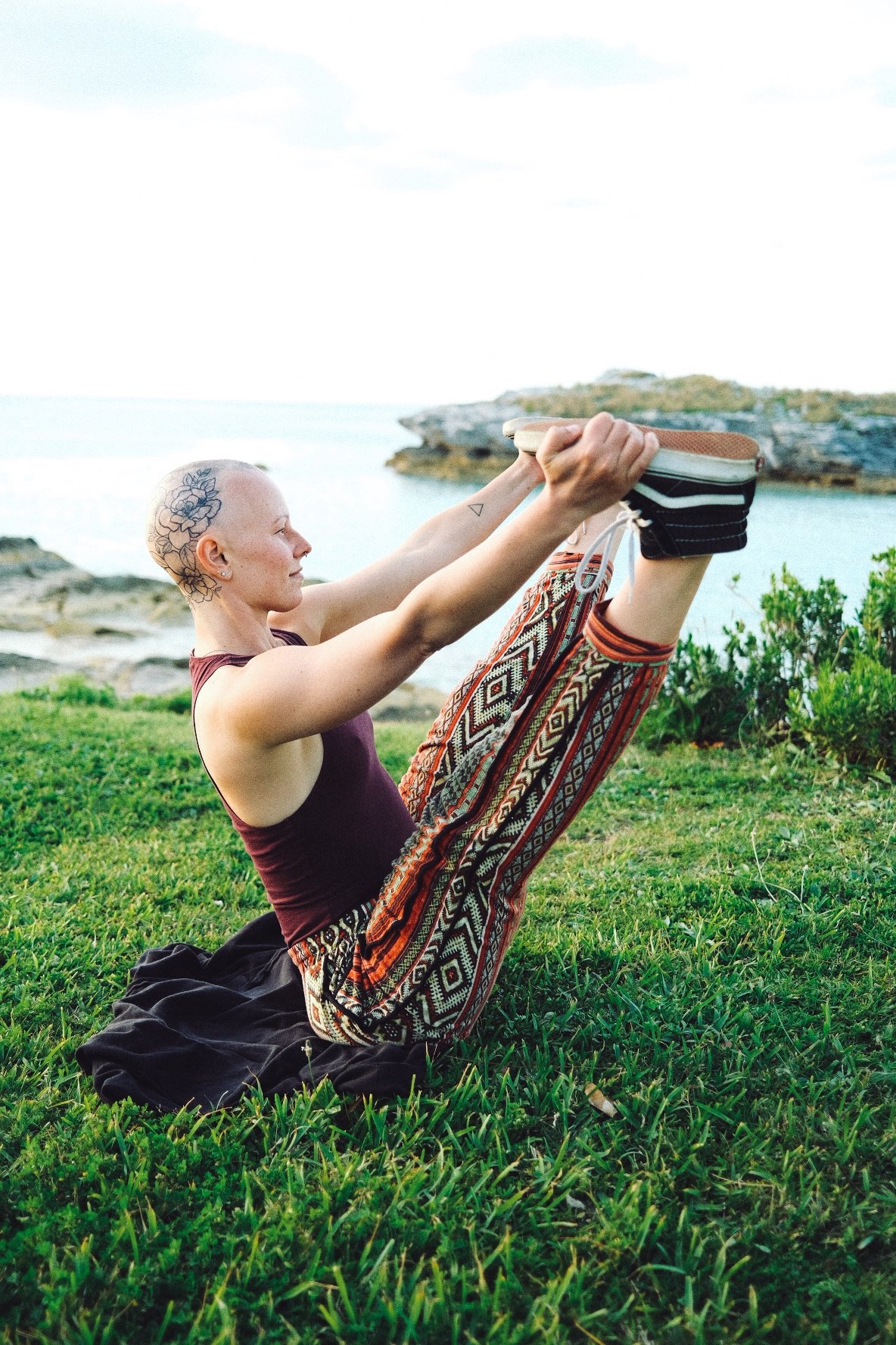 A woman with tattoos on her head and arm practicing yoga outdoors on grass near the water, holding her legs up in a seated forward bend pose.