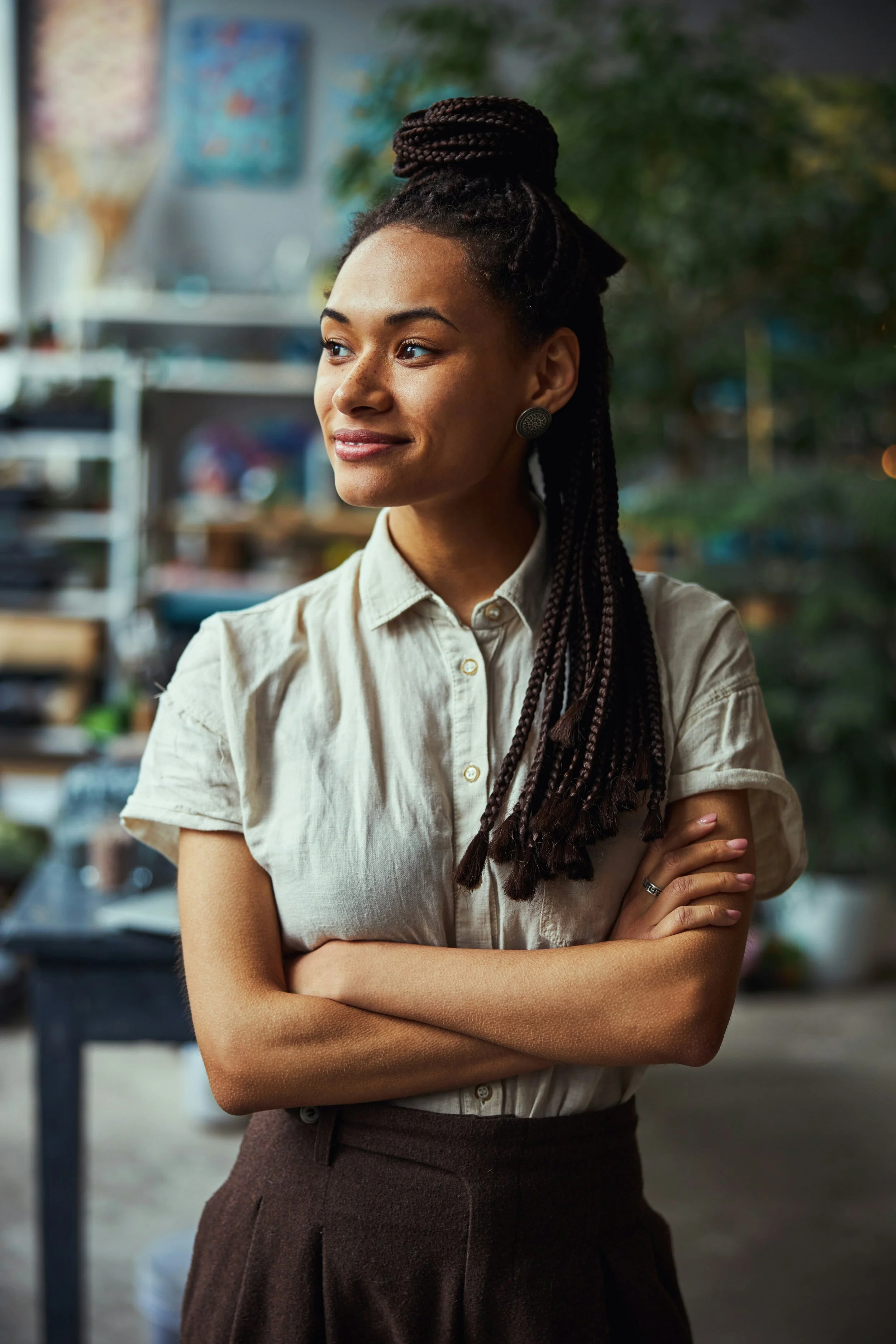 Jeune femme avec des cheveux en tresses et un chignon, portant une chemise beige, regardant sur le côté avec une expression calme et confiante, dans un intérieur avec des étagères et un décor flou en arrière-plan.