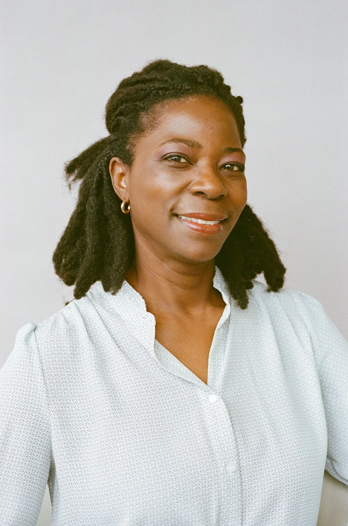 Une femme souriante portant une chemise à petits motifs, avec des cheveux bouclés en dreadlocks, portant des boucles d'oreilles en or, contre un fond neutre.