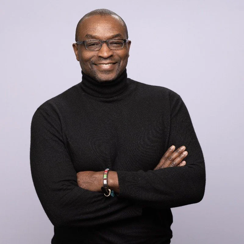 Homme souriant portant un chandail noir, avec des lunettes, bras croisés, fond gris clair.