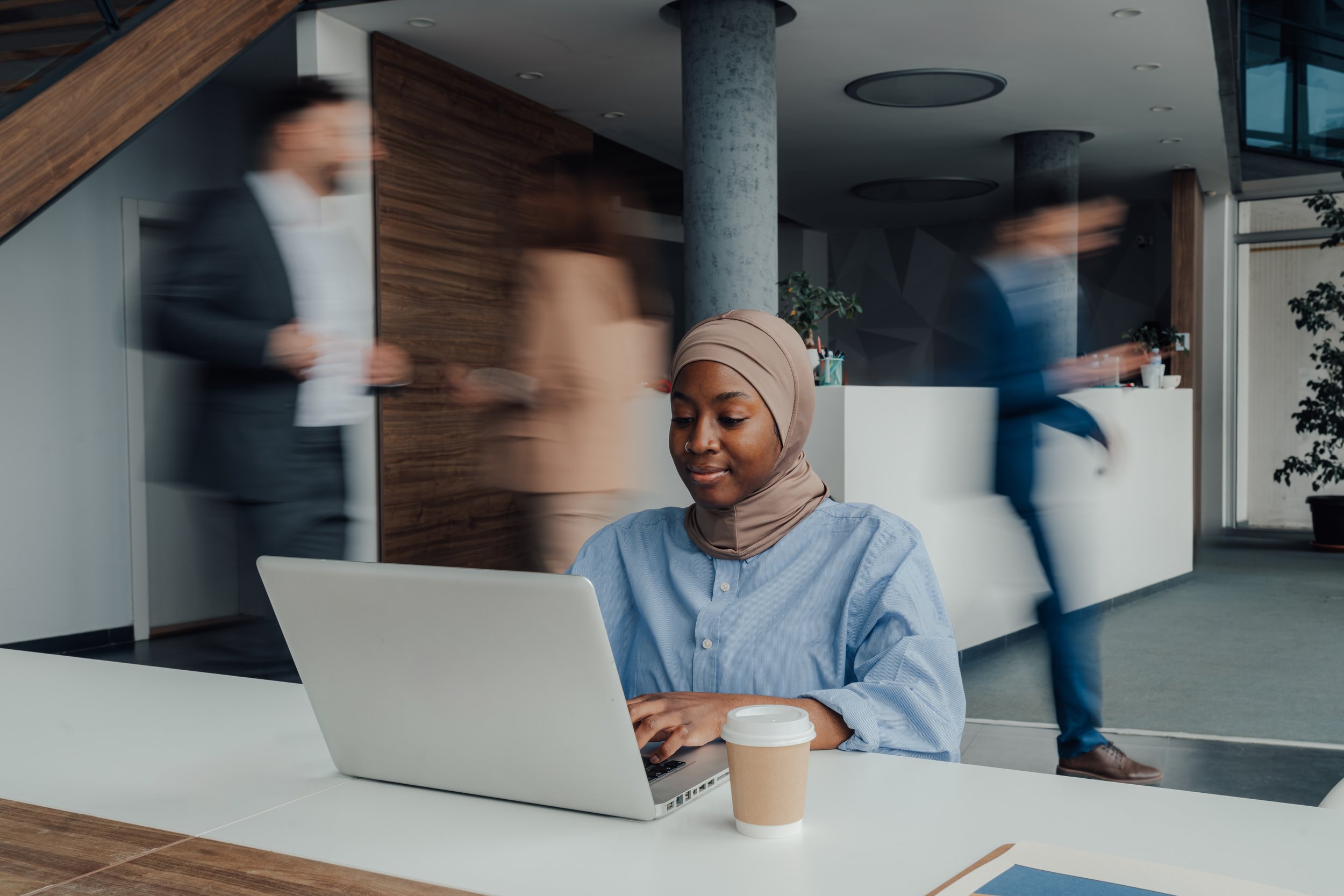 Une femme portant un hijab beige travaille sur un ordinateur portable à une table dans un espace de bureau moderne, avec un café à côté. Des personnes passent rapidement derrière elle, floues, dans un environnement intérieur avec des colonnes en béton et des murs en bois.