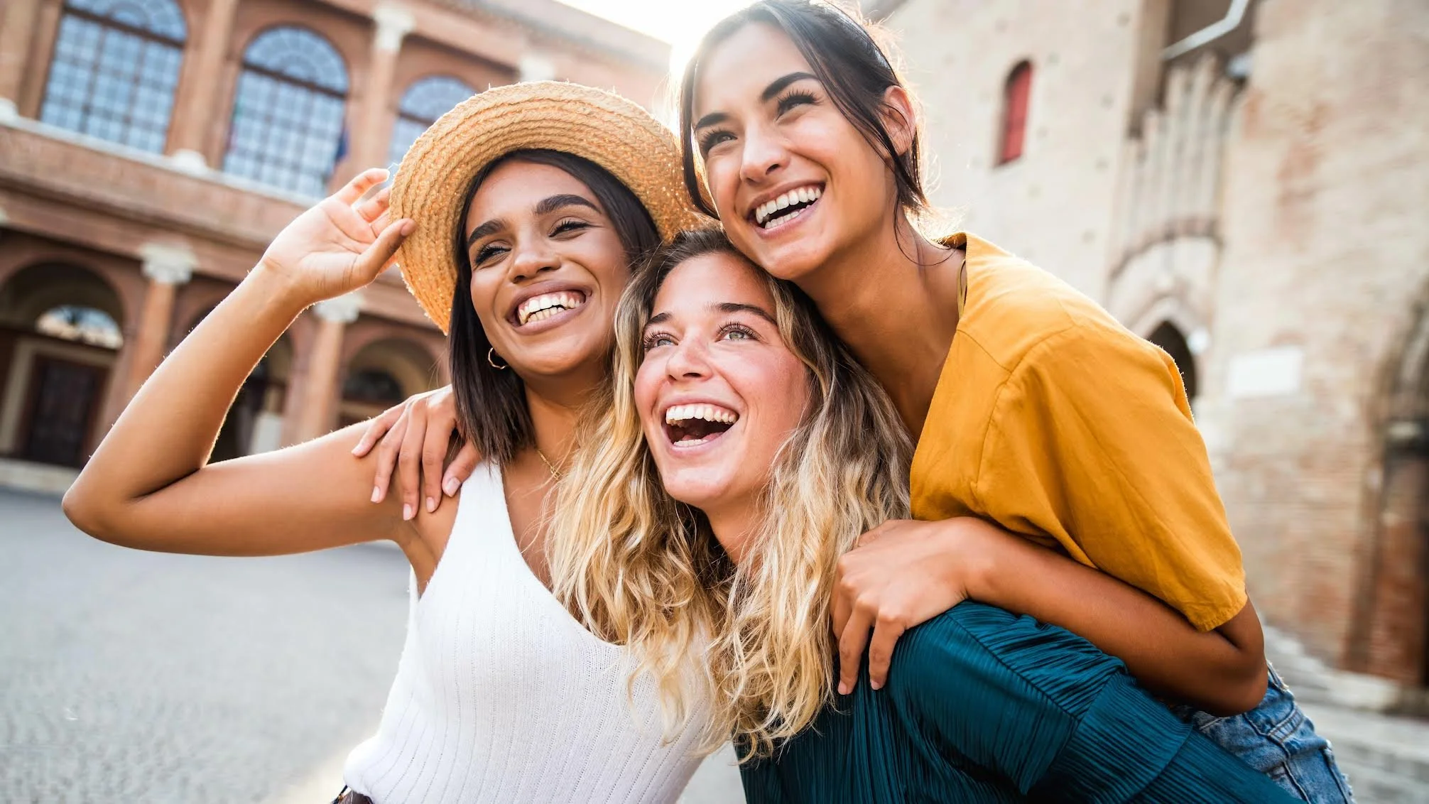 Three young women smiling and hugging each other outdoors on a sunny day, with historic brick buildings in the background.