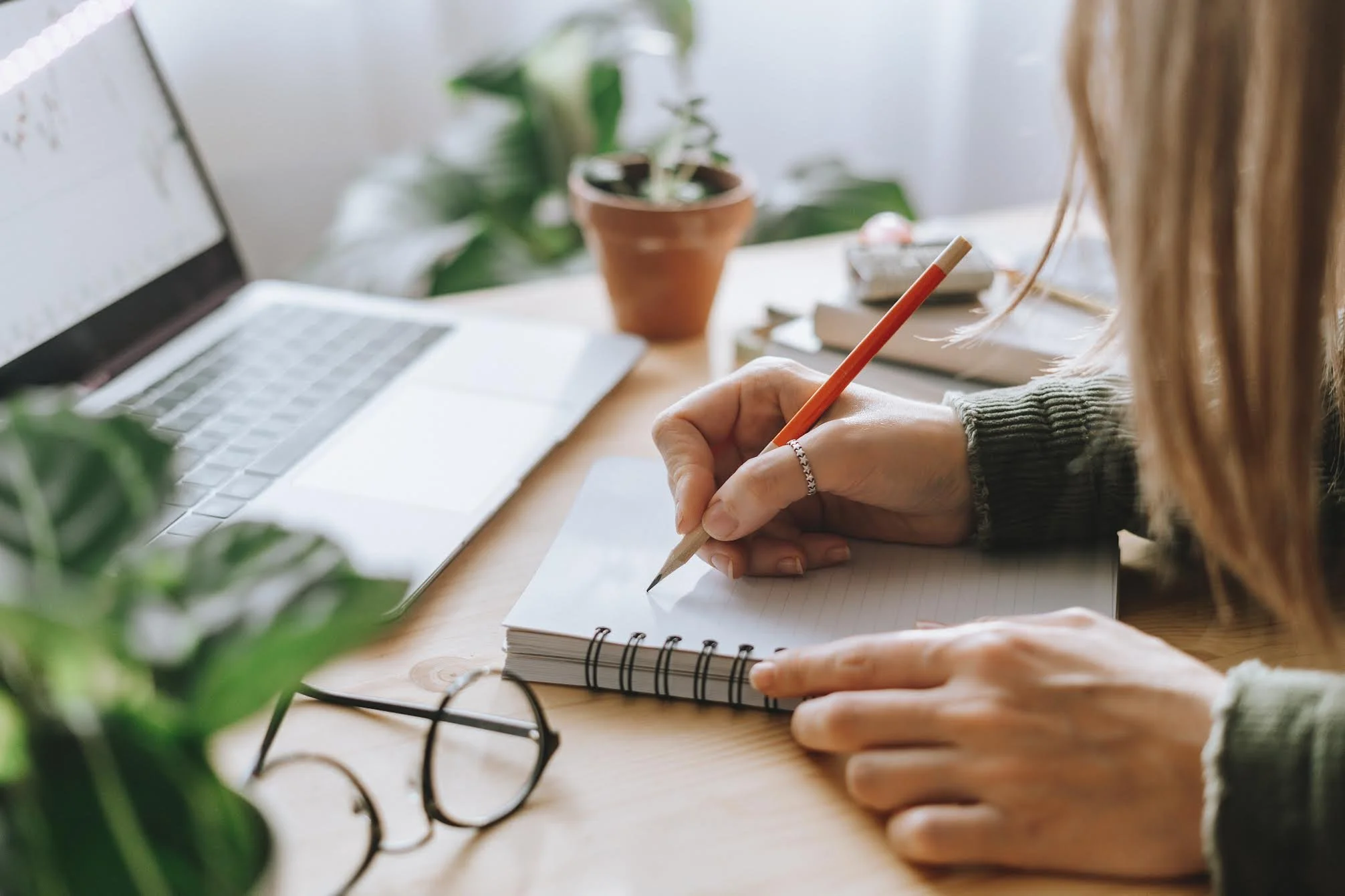 Person writing in a blank spiral notebook with a pencil, on a wooden desk with a laptop, potted plant, and glasses.