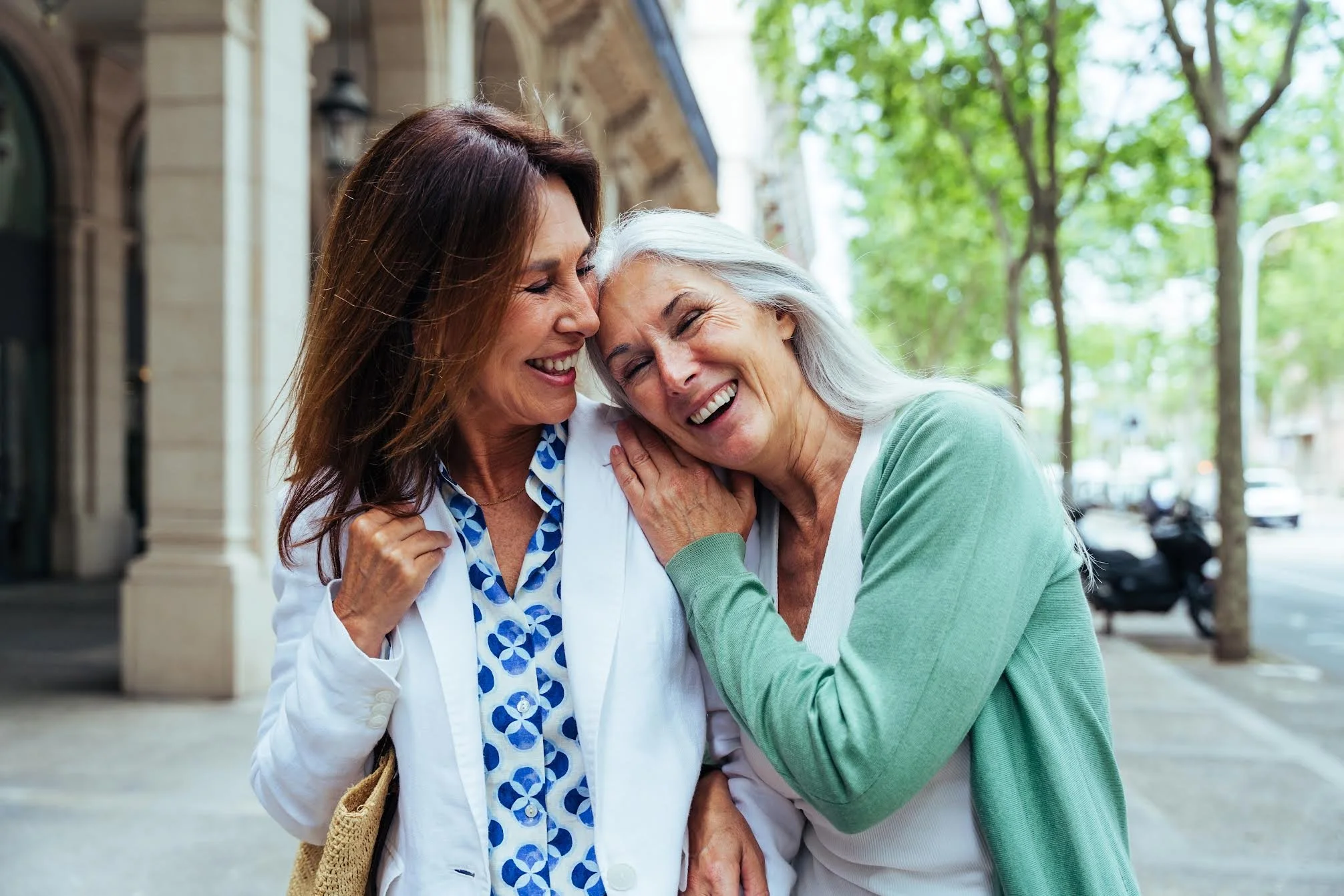 Two middle-aged women share a hug and laugh together outdoors on a city street with trees and buildings in the background.