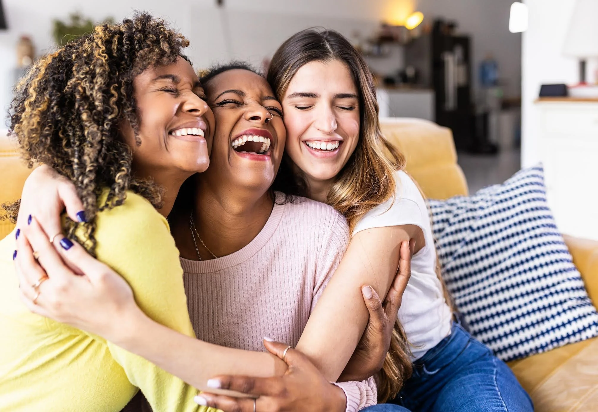 Three women hugging and smiling happily in a cozy living room.