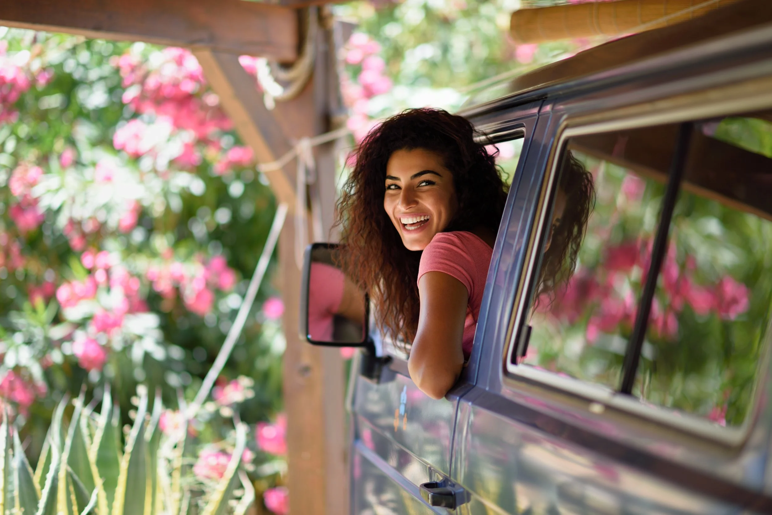 Woman with curly dark hair smiling and leaning out of a vehicle window, surrounded by pink flowers and green foliage.