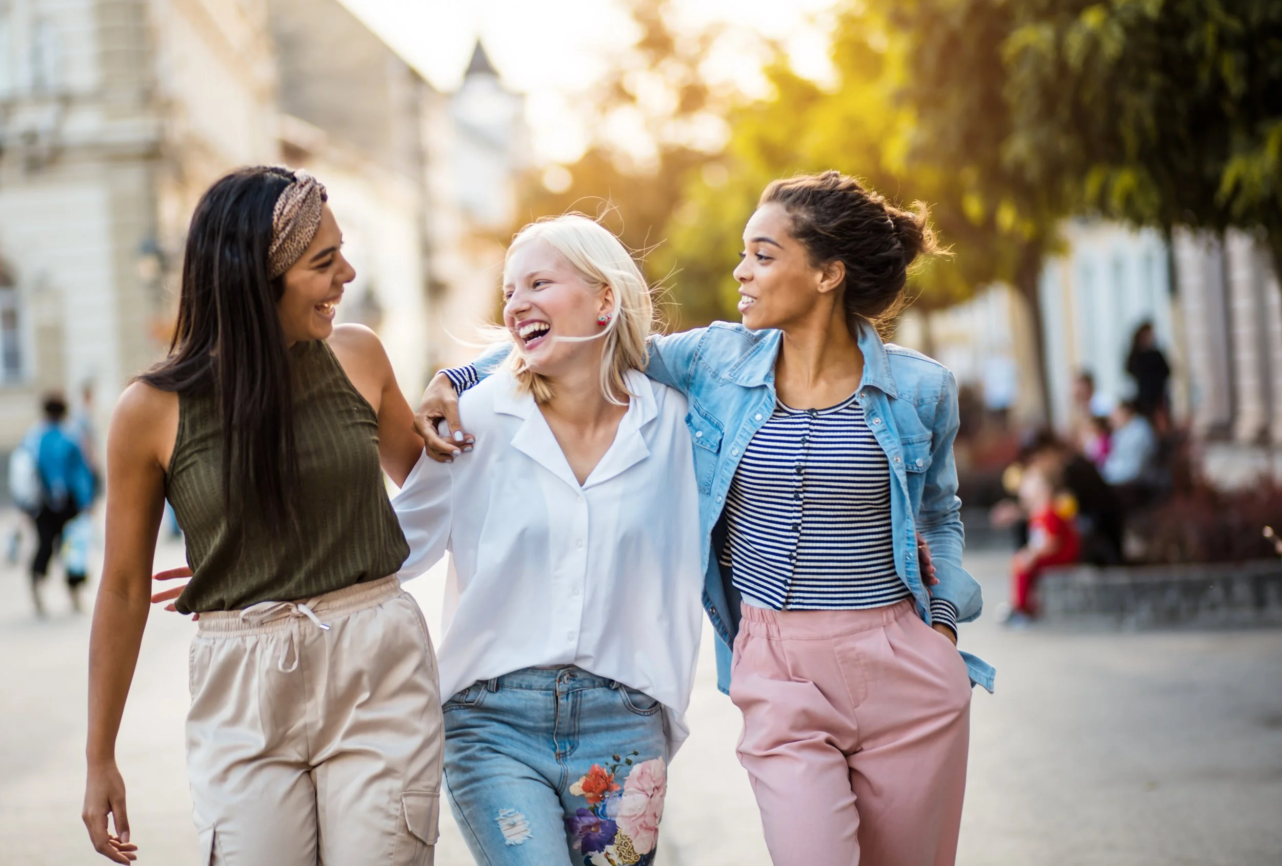 Three women walk arm-in-arm outdoors in a city park on a sunny day, smiling and laughing, with blurred trees and buildings in the background.