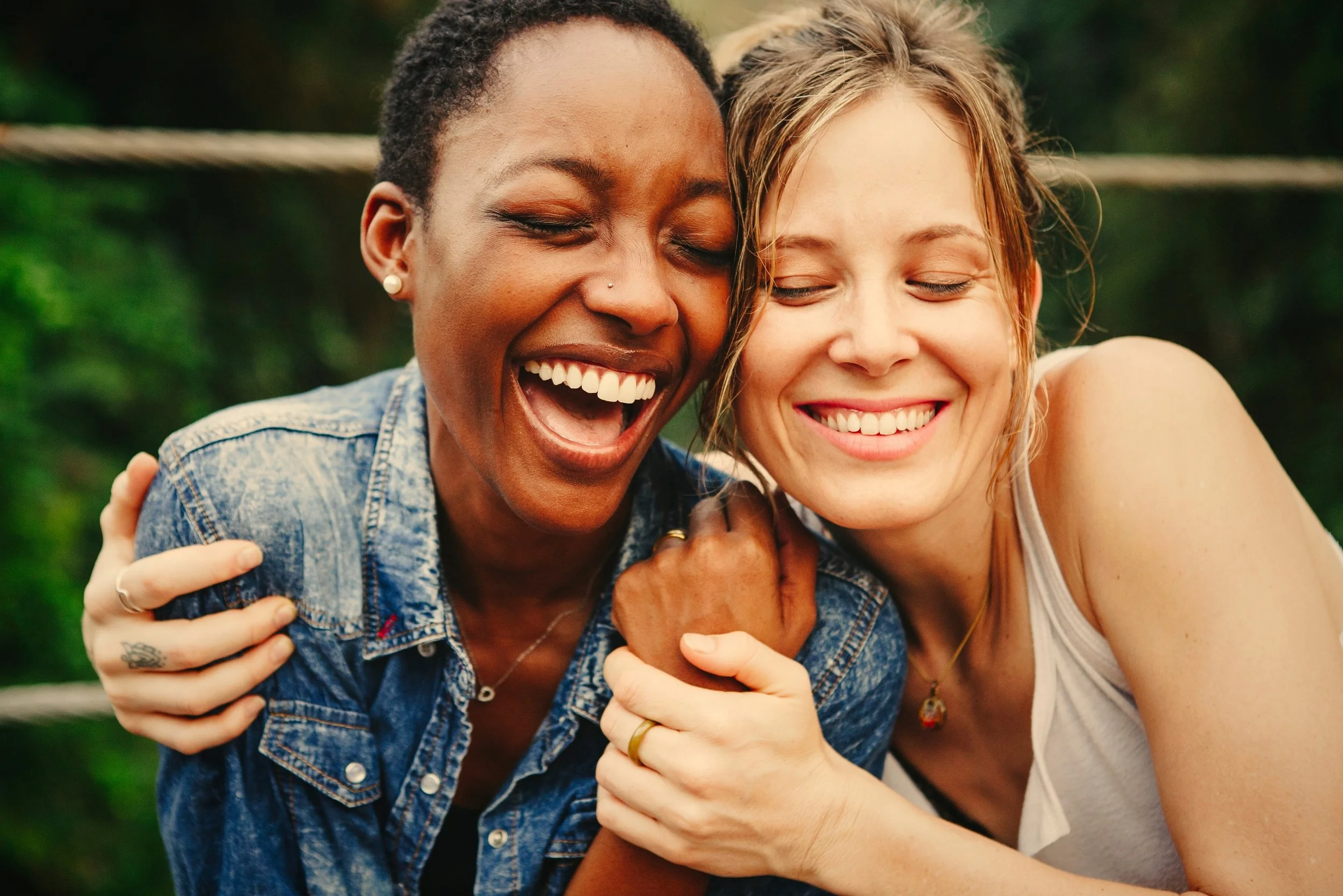 Two women smiling and hugging outdoors, with green blurred background.