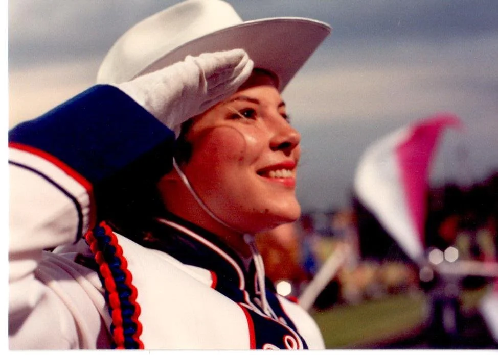 A young woman in a band uniform saluting with her right hand, wearing a white cowboy hat and white gloves, in an outdoor setting during dusk or evening.