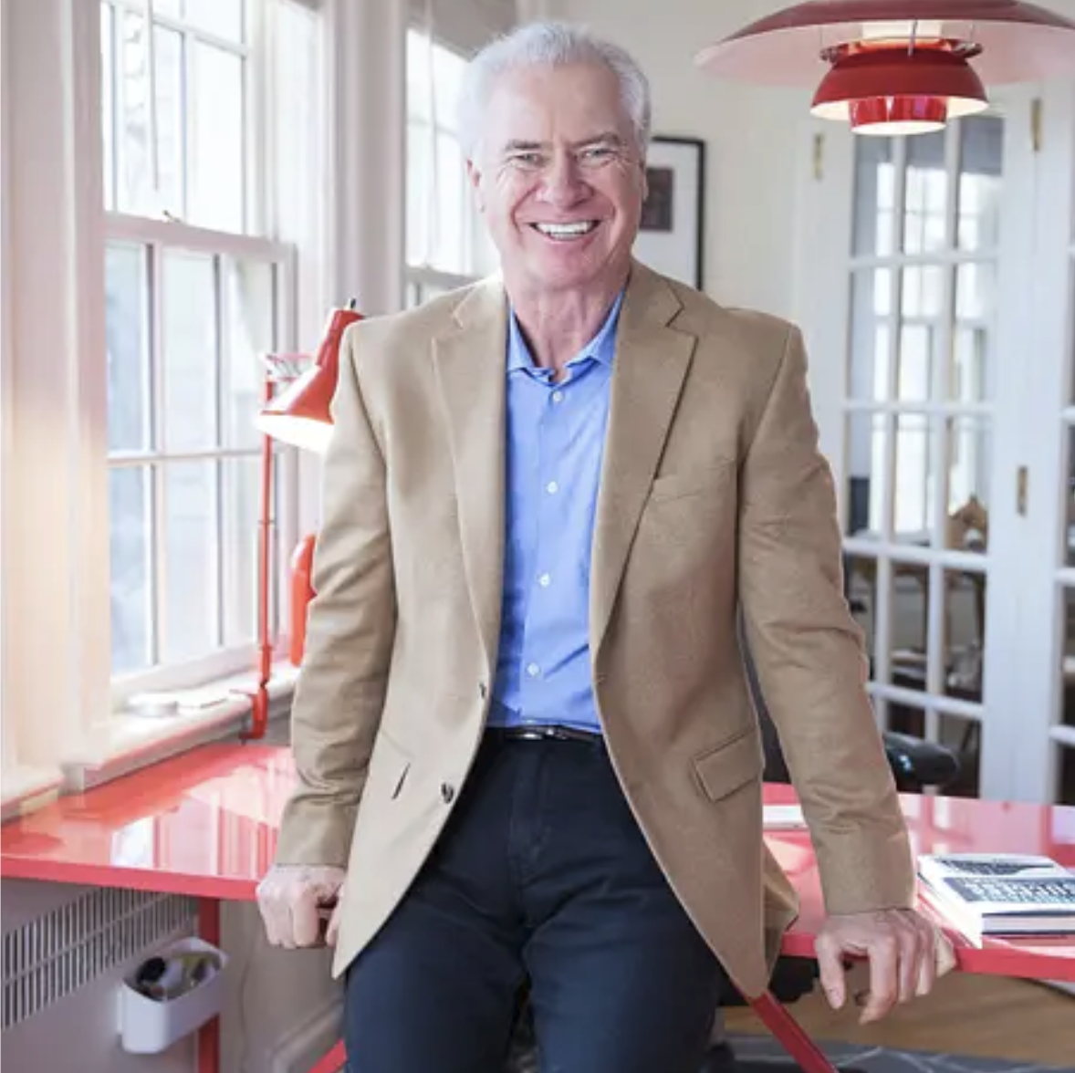 A smiling elderly man with white hair, wearing a tan blazer and blue shirt, leaning on a red table in a bright room with large windows.
