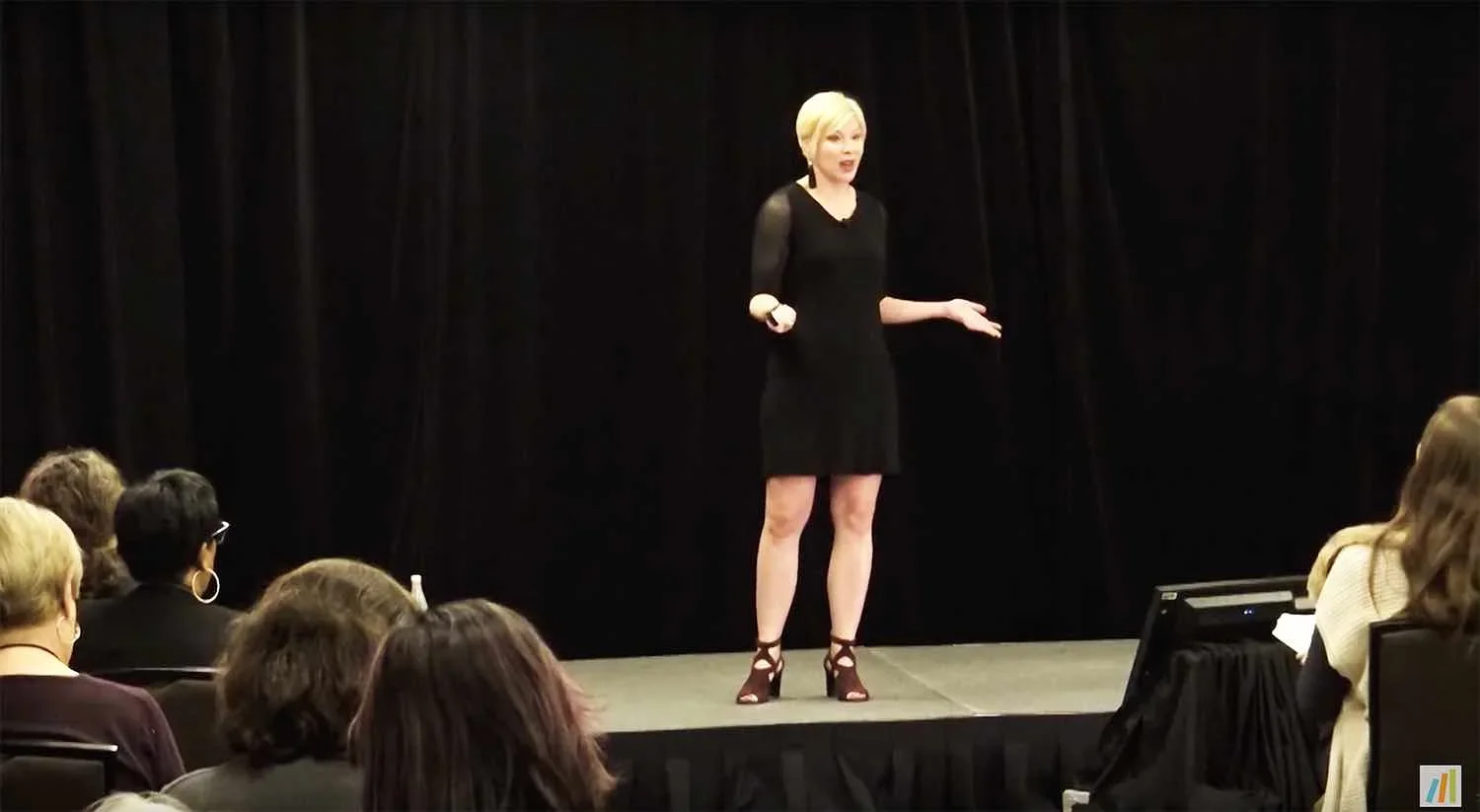 A woman wearing a black dress and high heels is standing on a stage, speaking to an audience while holding a microphone.