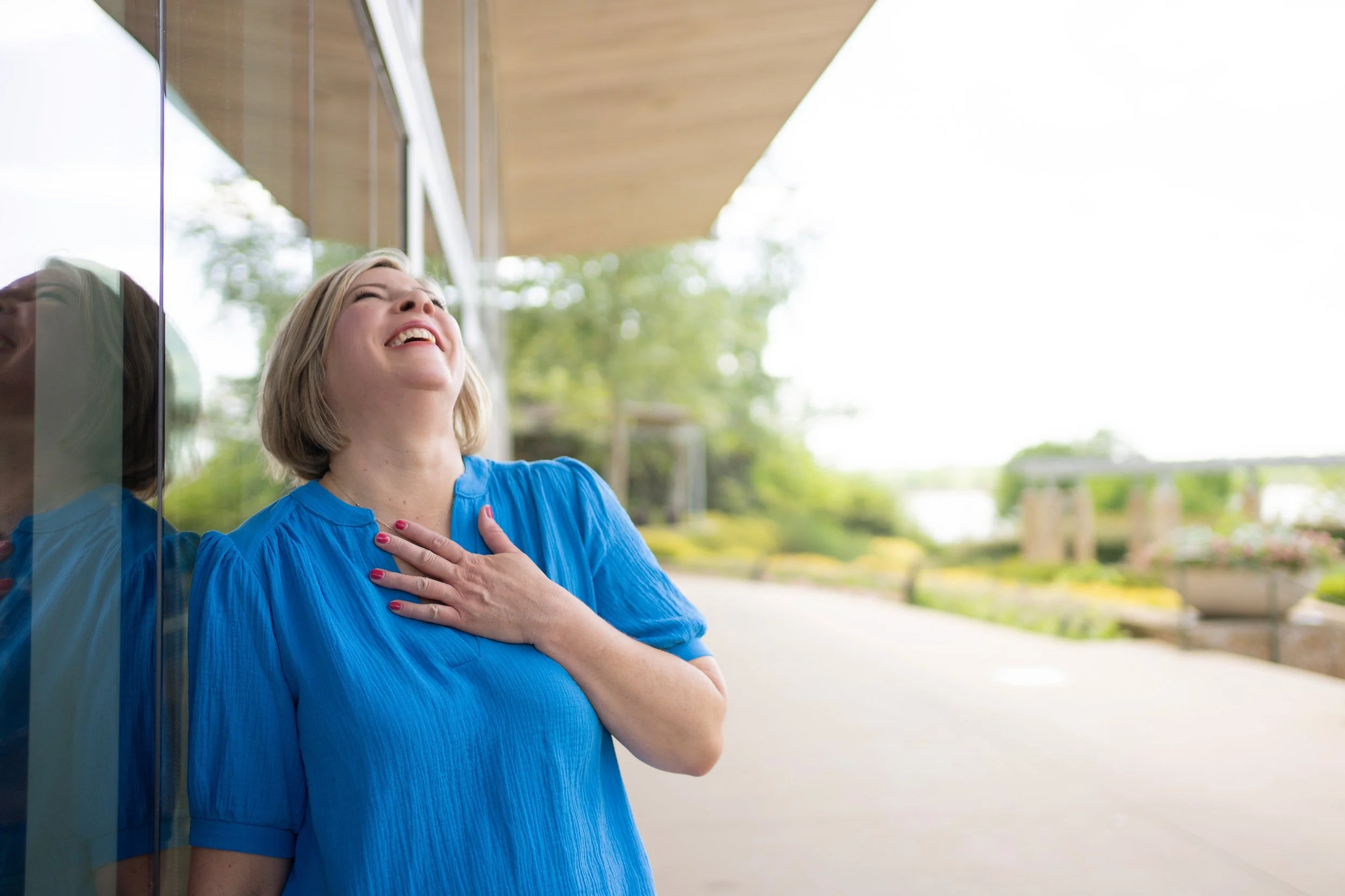 A woman with blonde hair in a blue dress is leaning against a reflective glass wall, holding her chest and laughing outdoors.