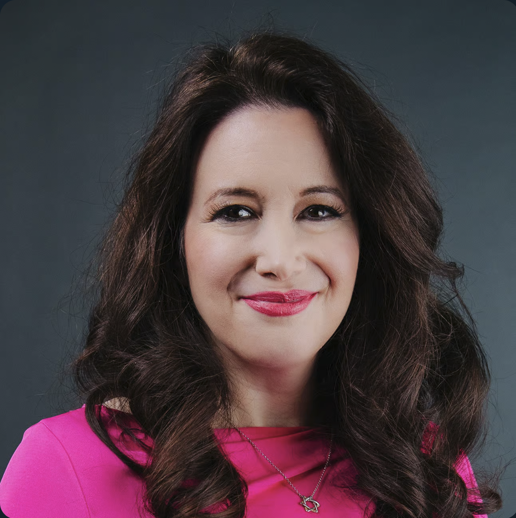 A woman with long, dark, wavy hair wearing a pink top and a necklace, smiling against a dark background.