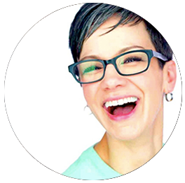 Close-up of a woman with short dark hair, glasses, and earrings, smiling and laughing.