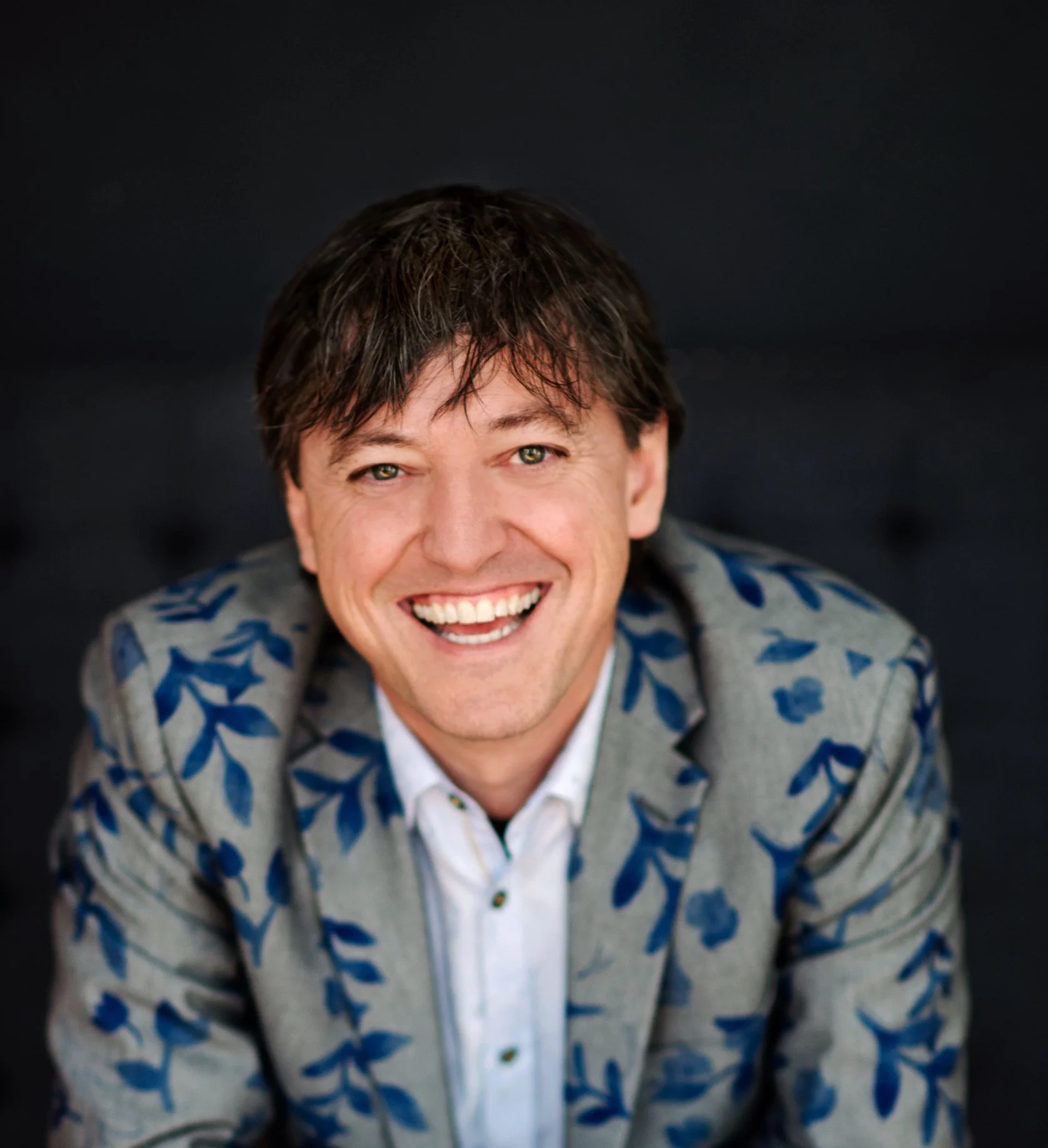 Man smiling, wearing a grey jacket with blue floral pattern and a white shirt, against a dark background.