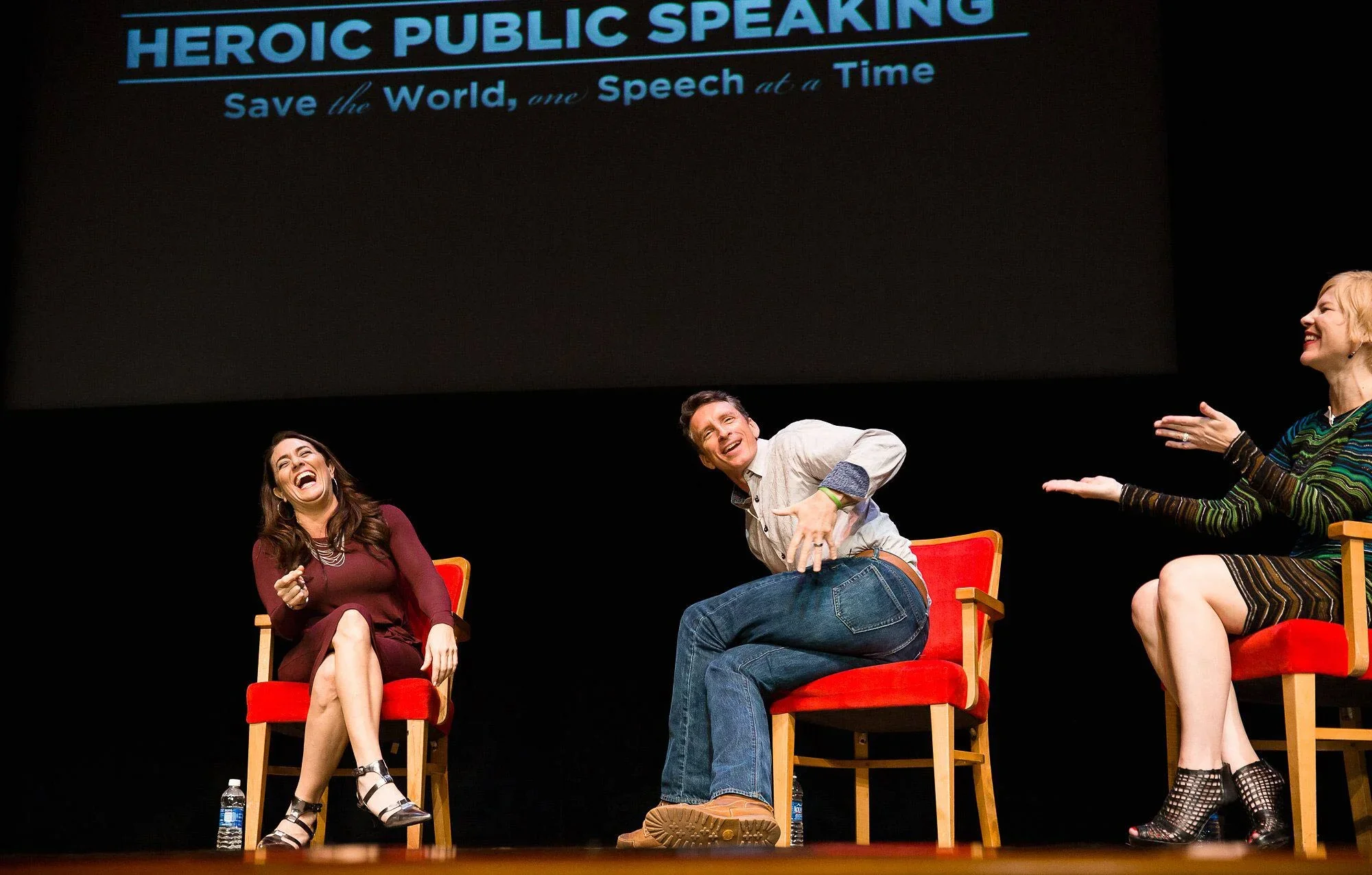 Three people are sitting on red chairs on stage, laughing and engaging in conversation during a panel discussion or presentation titled "Heroic Public Speaking." The background shows a large screen with the event's theme and slogan: "Save the World, one Speech at a Time."