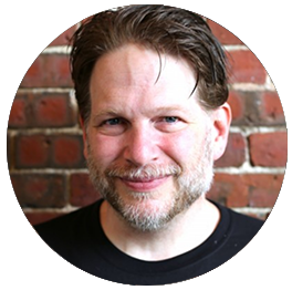Chris Brogan with brown hair, a beard, and a mustache, smiling, in front of a brick wall.
