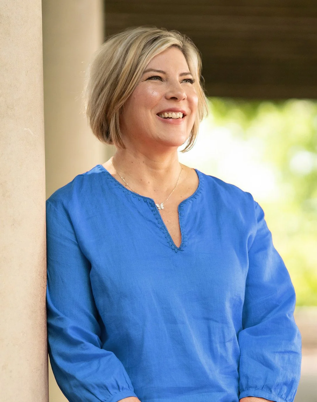 A woman with short blonde hair wearing a blue top, smiling and leaning against a beige wall outdoors with green trees in the background.