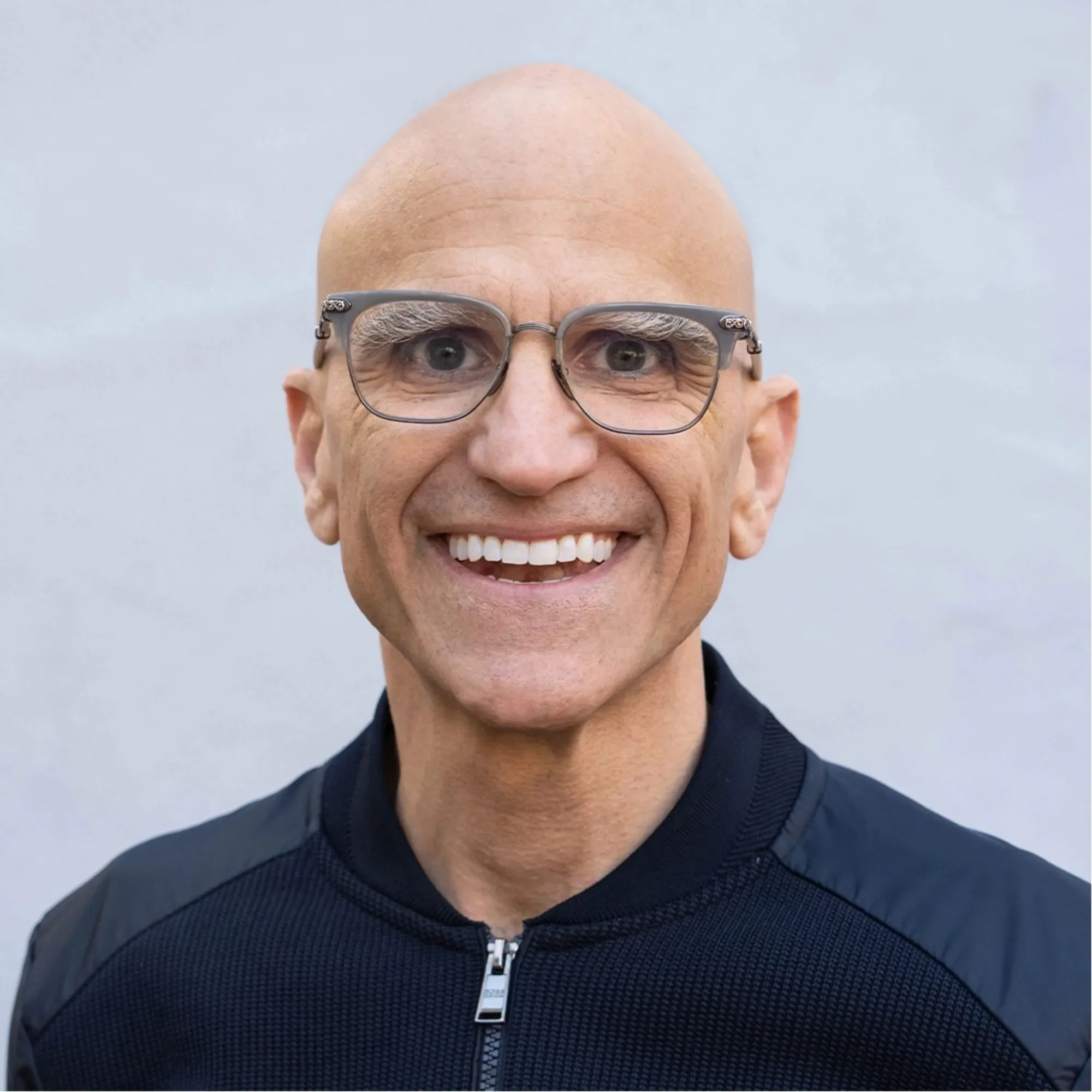 Close-up of a smiling man with glasses and a bald head, wearing a black zip-up jacket, against a plain gray background.