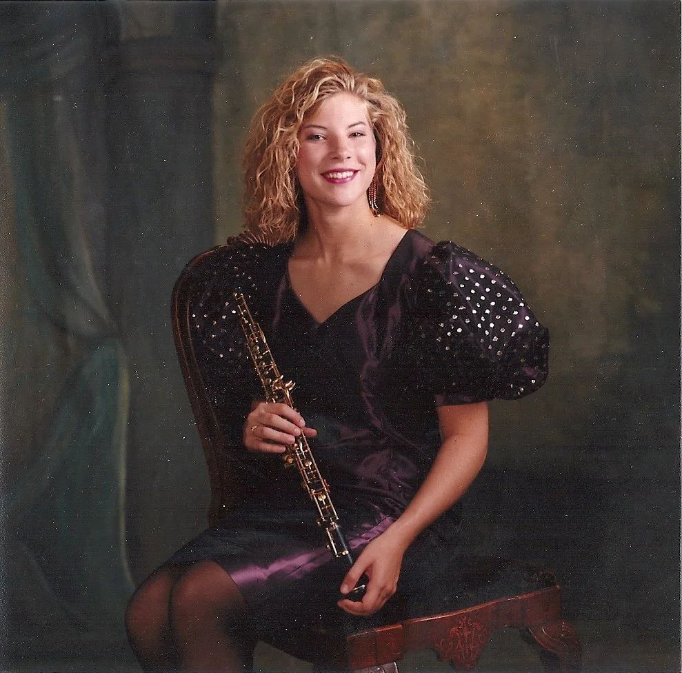 A woman with curly blonde hair wearing a black dress with shiny, puffy sleeves, sitting on a wooden stool, holding a flute, and smiling at the camera against a dark, textured background.